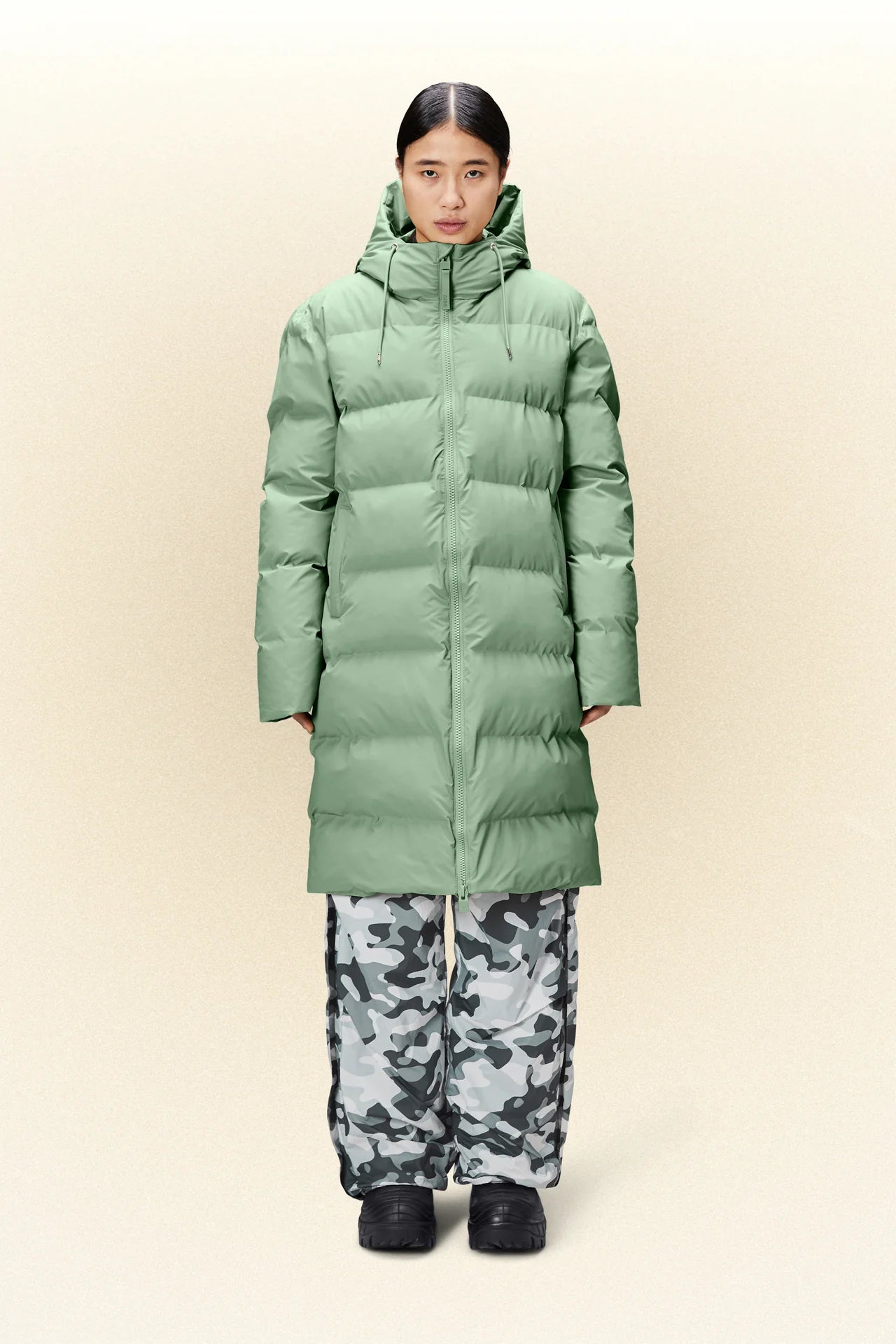 A woman wearing a green Alta Long Puffer Jacket, a waterproof and winter jacket by Rains, paired with camouflage pants.