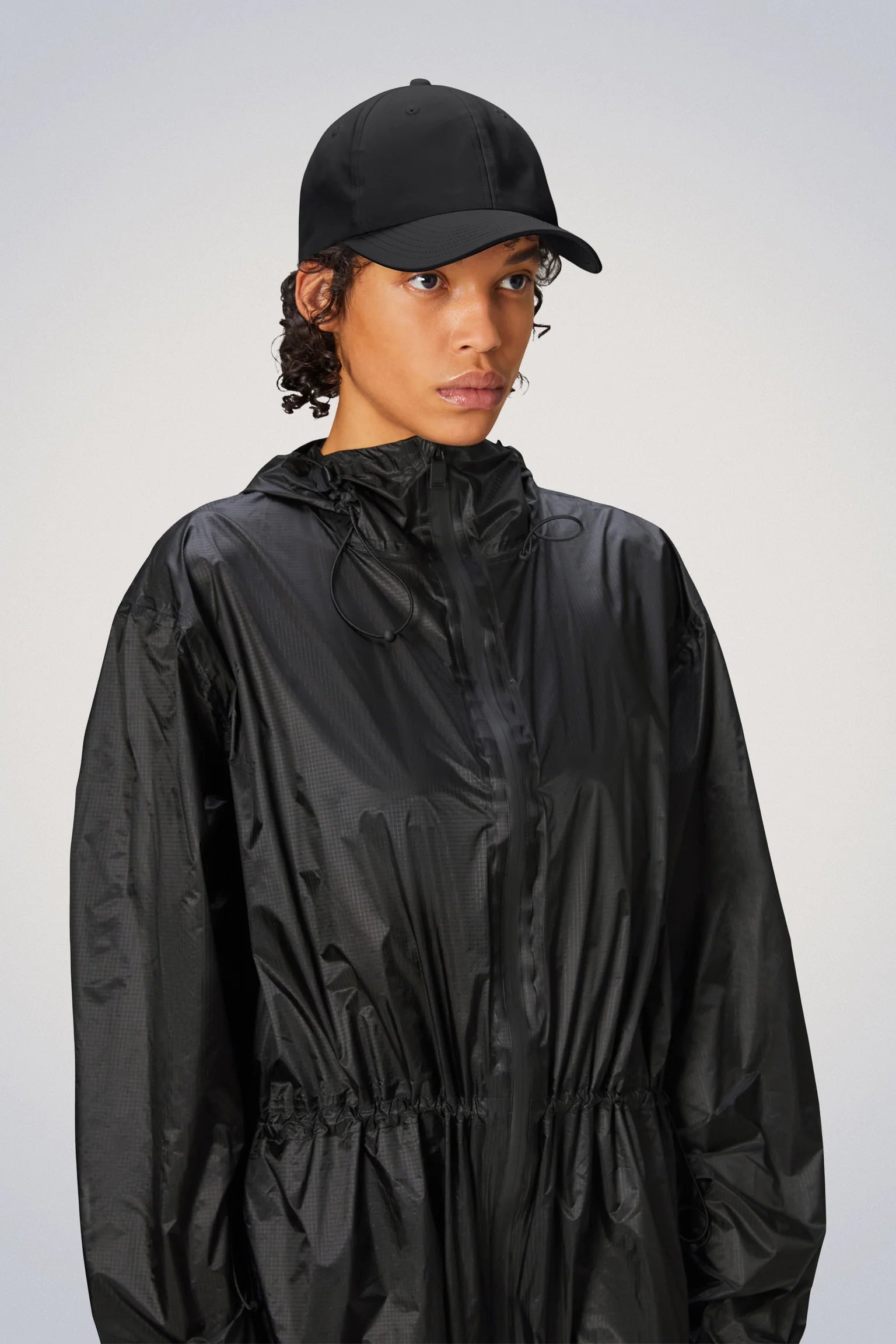 A woman wearing a black raincoat and hat with a Rains Waterproof Cap.