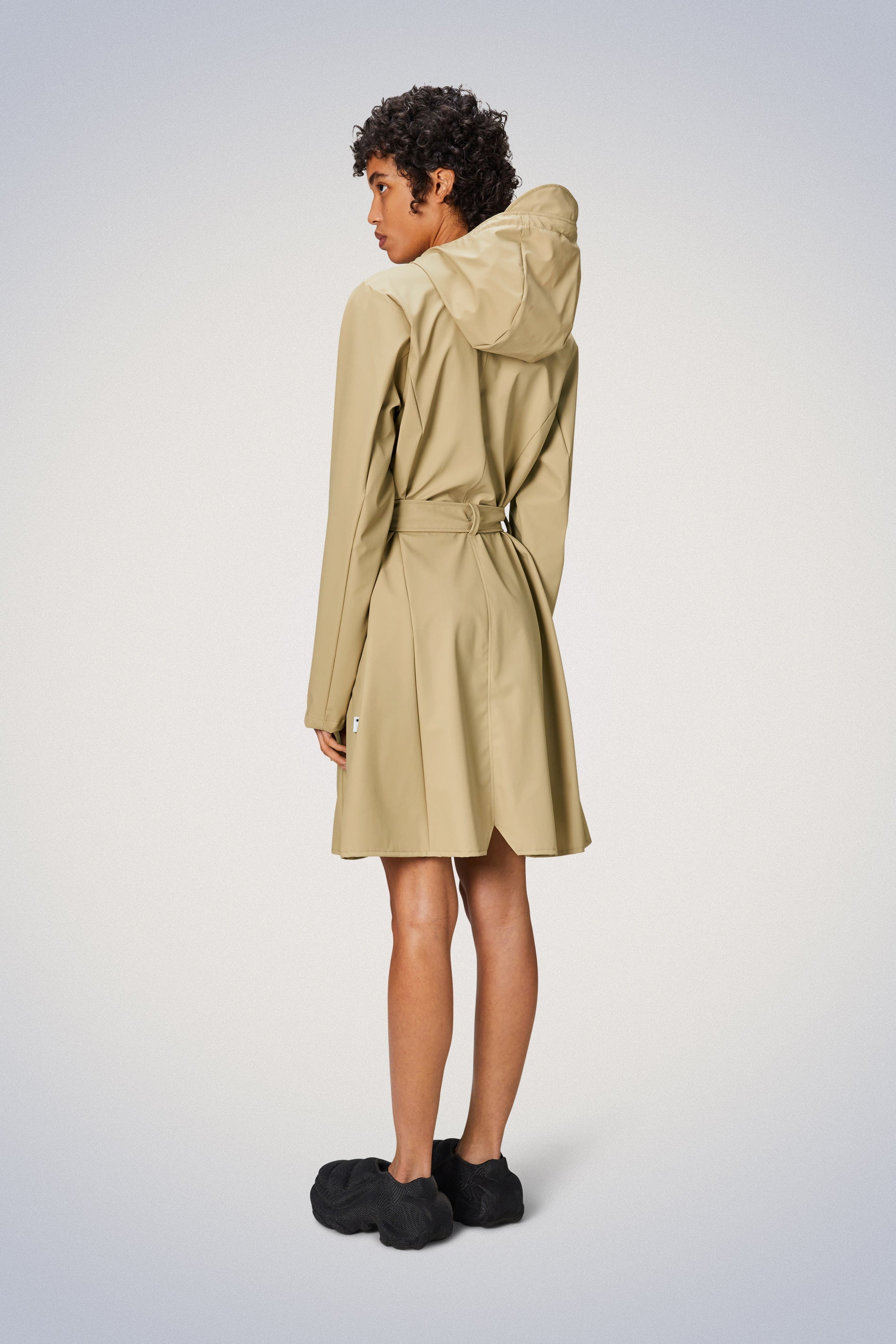 The Curve Jacket - Sand by Rains is a beige trench coat that combines practicality and style. It features a double-breasted front, water-resistant material, and a back vent for ease of movement.