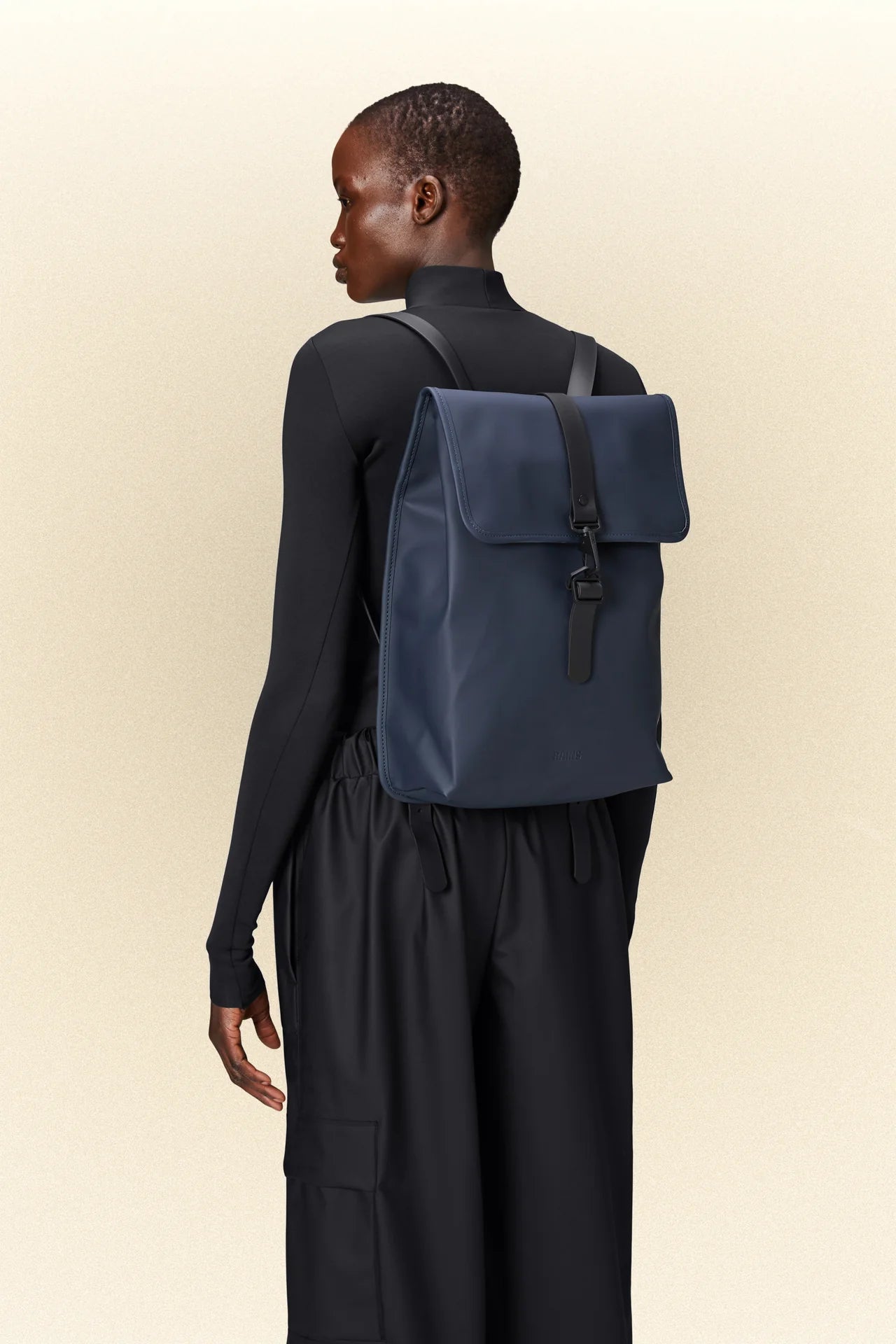 The back of a woman wearing black pants and a blue Rains waterproof rucksack.