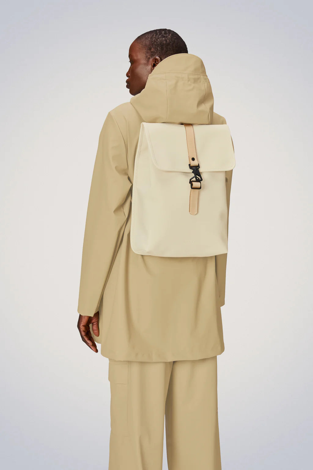 Rains Rucksack: The back view of a woman wearing a beige Rains rucksack, this perfect companion includes a laptop sleeve and two side pockets for easy access to water bottles or other essentials. The padded