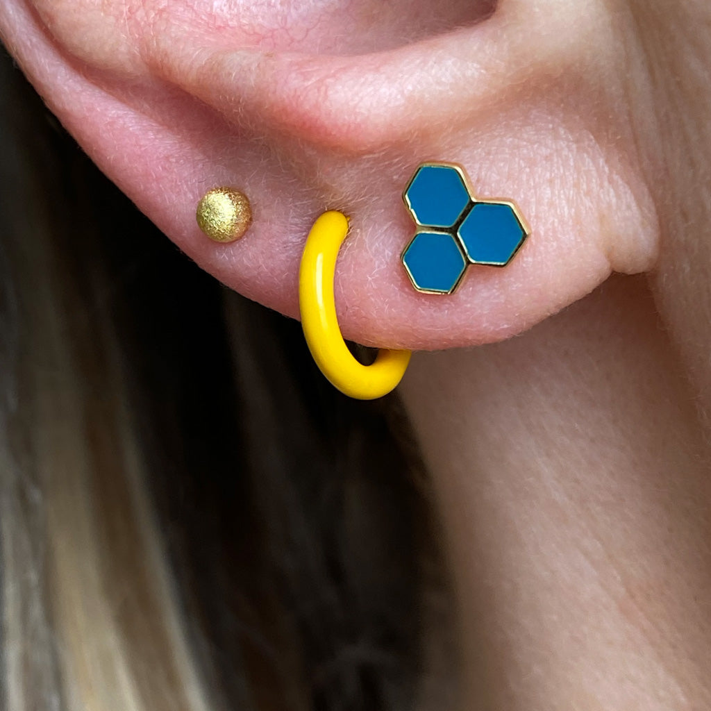Close-up of a woman's ear showing three stacking earrings: a Brushed Ball Single Stud - Gold by Lulu Copenhagen, a bright yellow hoop, and a blue hexagonal stud.