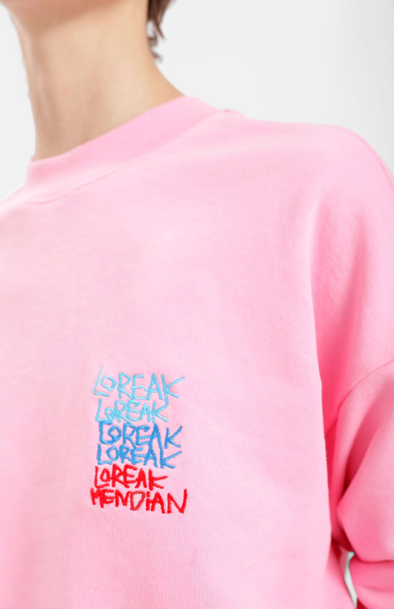 Person wearing a pink shirt with a repeated embroidered design of the words "L'oreak" in blue, light blue, and red.
