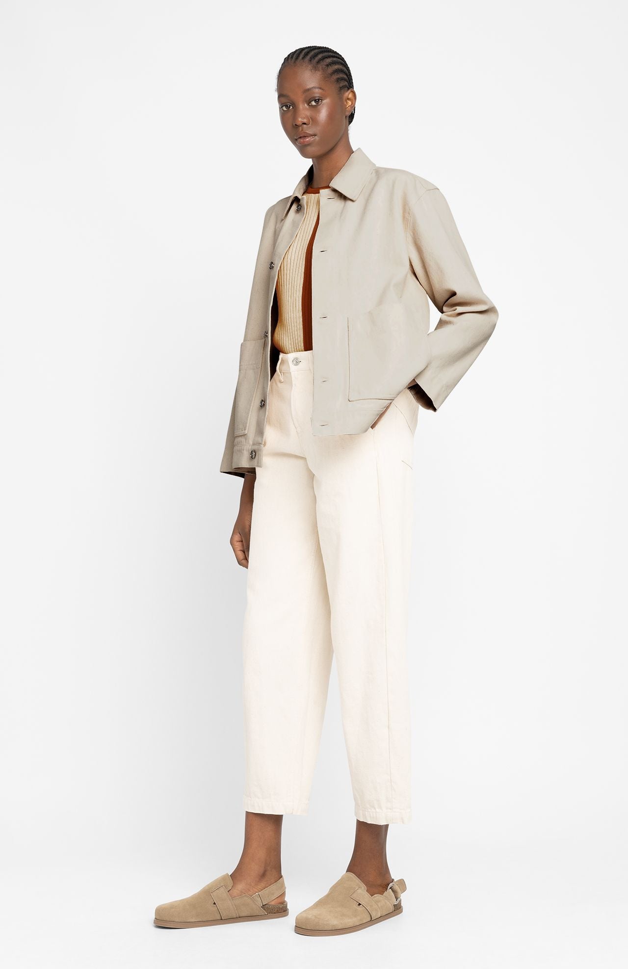 A woman stands against a plain background wearing a light tan jacket, ribbed brown top, cream high-waisted pants, and beige loafers.