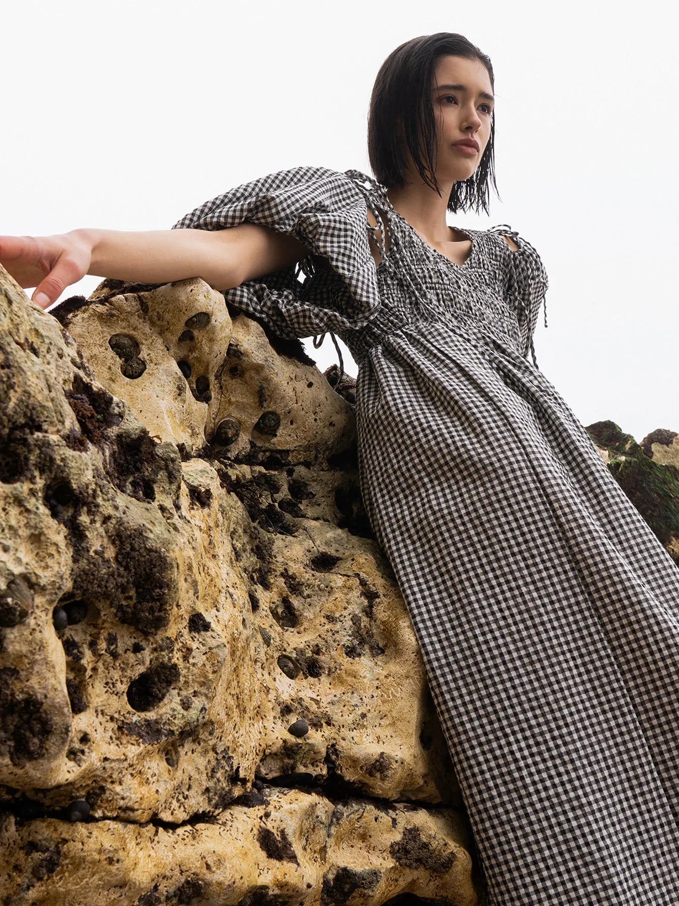 A person wearing a loose-fitting white blouse and beige skirt stands against a dark background, looking towards the camera.