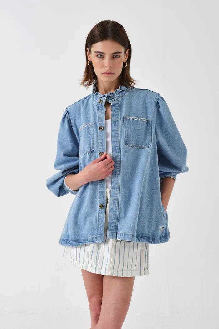 Woman wearing a light blue denim jacket over a white dress with blue vertical stripes, standing against a plain white background.