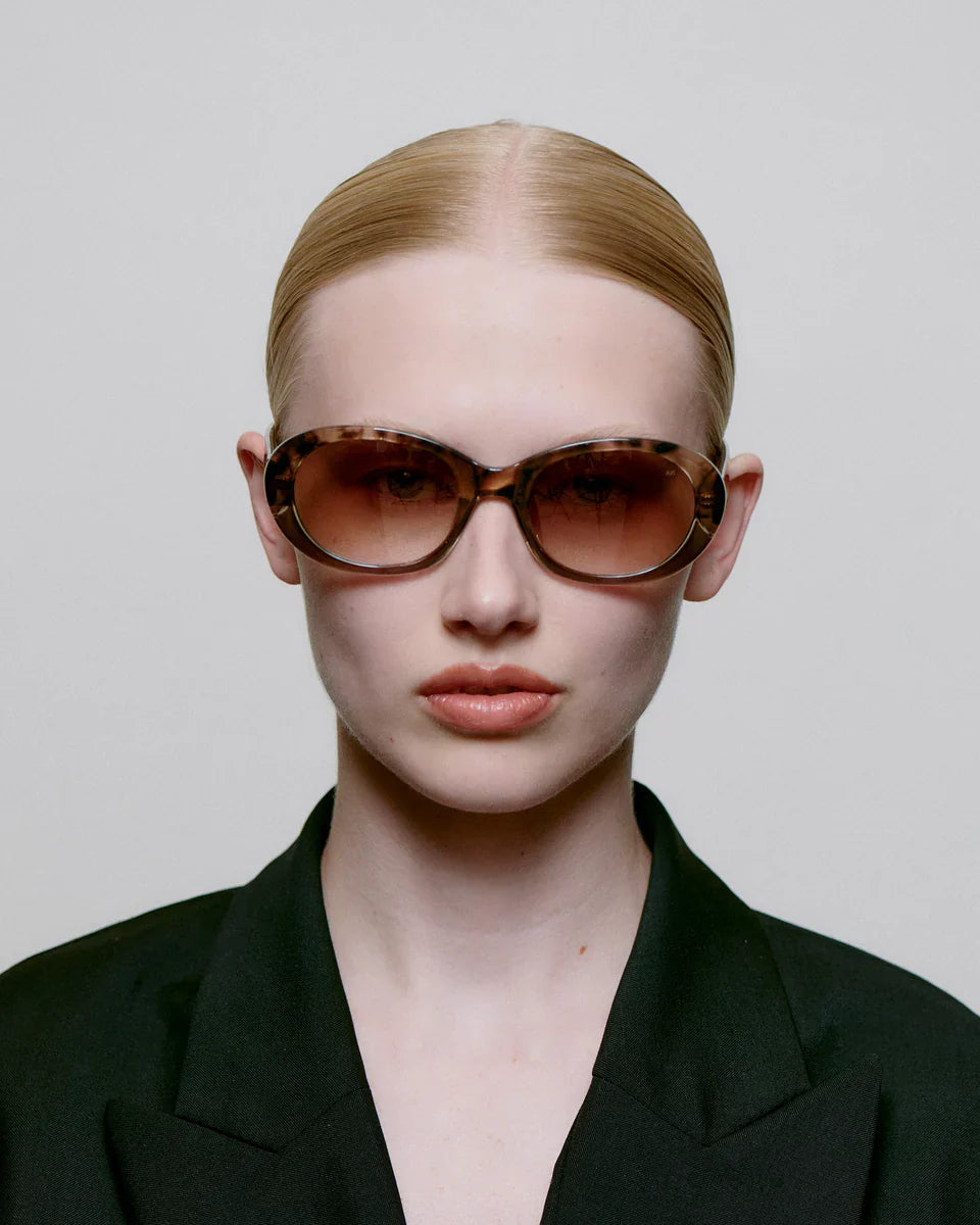 A person with slicked-back blonde hair wearing large, oval tortoiseshell sunglasses and a black blazer, facing forward against a plain background.