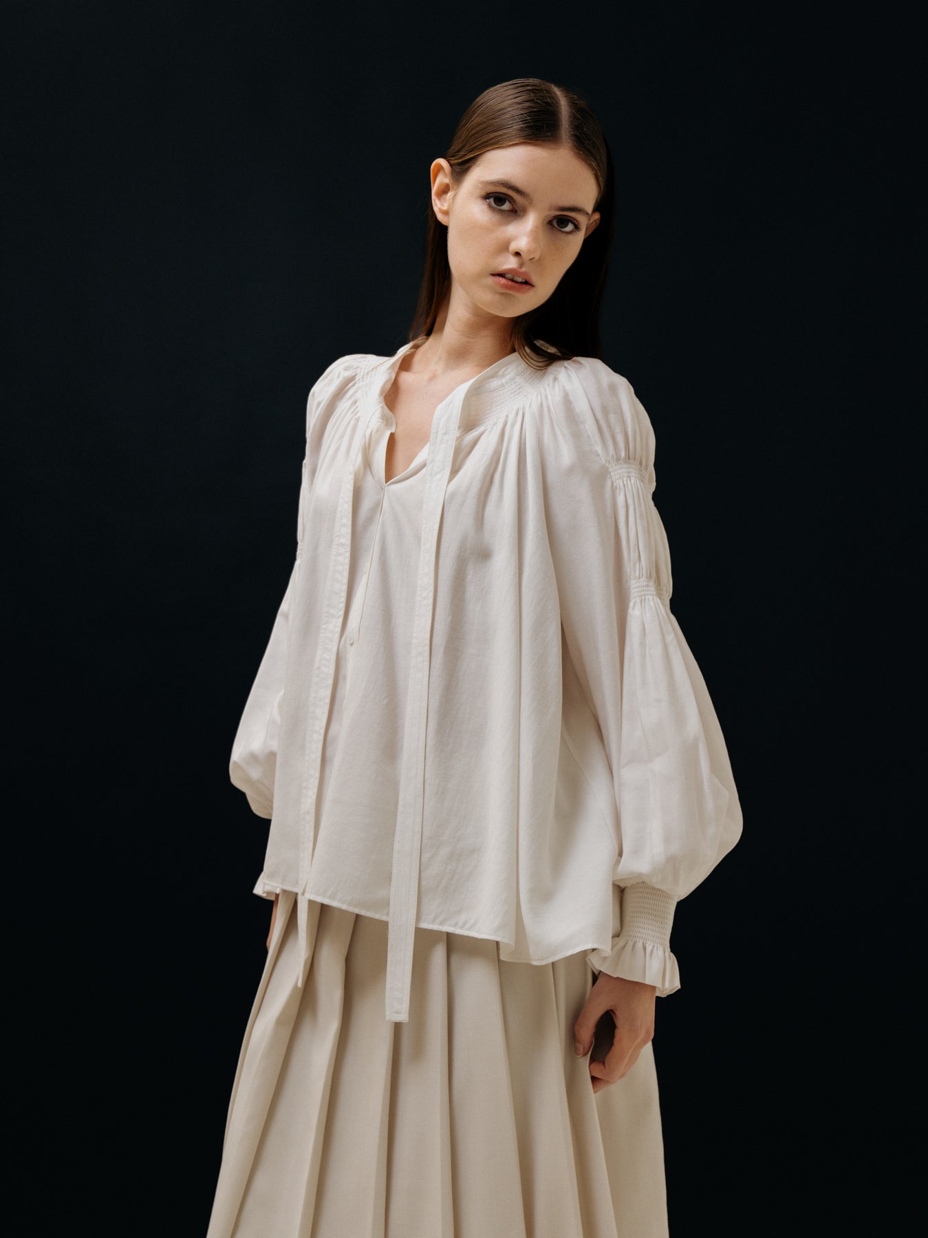 A person wearing a loose-fitting white blouse and beige skirt stands against a dark background, looking towards the camera.