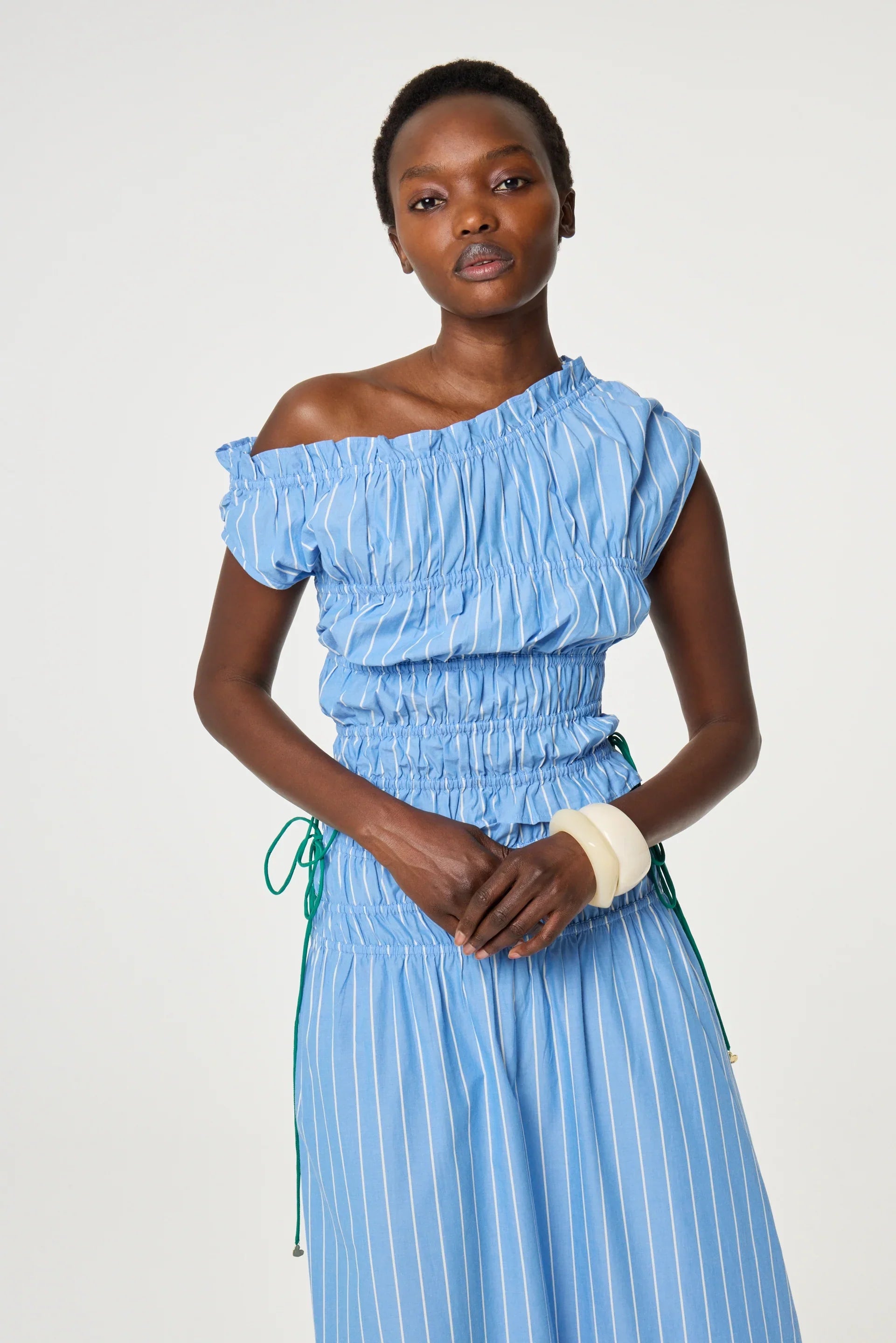A woman wearing a blue, one-shoulder, pinstriped dress with ruched detailing stands against a plain background.