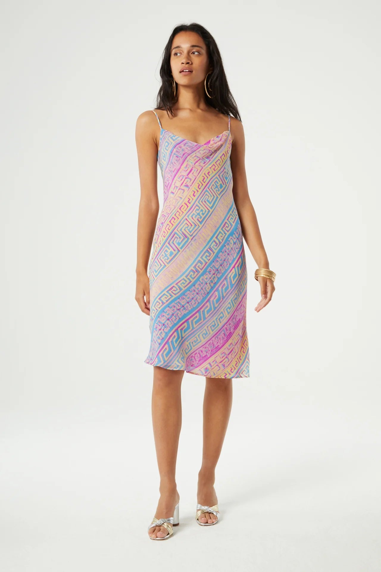 A woman stands wearing a multicolored, geometric-patterned slip dress with thin straps. She has long hair and is accessorized with gold bracelets, rings, and transparent high-heeled sandals.