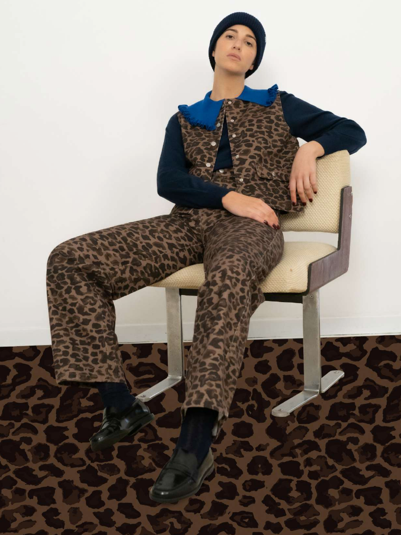 Person sitting on a beige chair, wearing a navy beanie, a blue-collared top, and matching brown leopard print vest and pants with black shoes; leopard print floor.