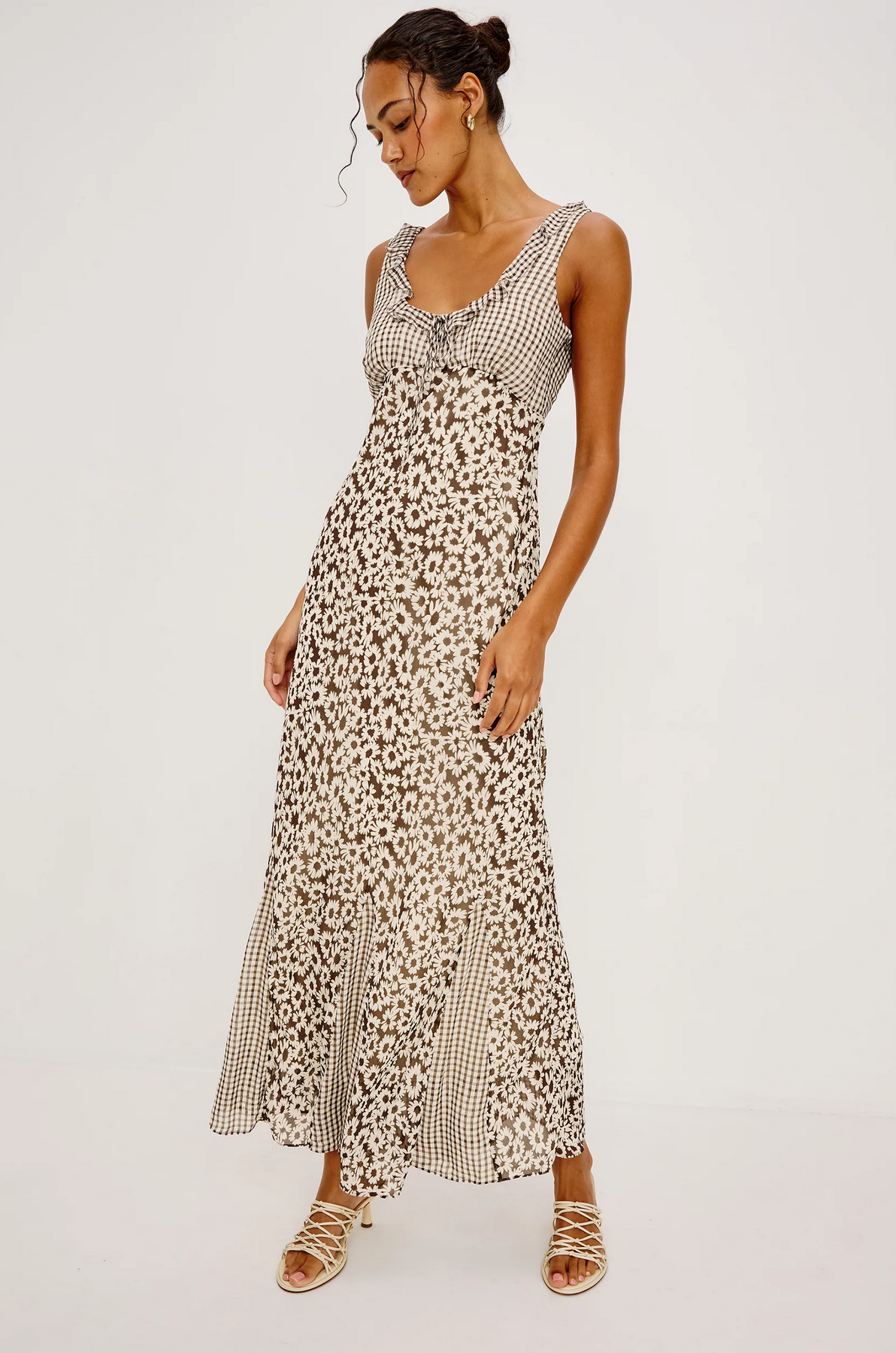 Woman wearing a sleeveless, ankle-length dress with a brown floral and checkered pattern, looking down and standing against a plain white background.