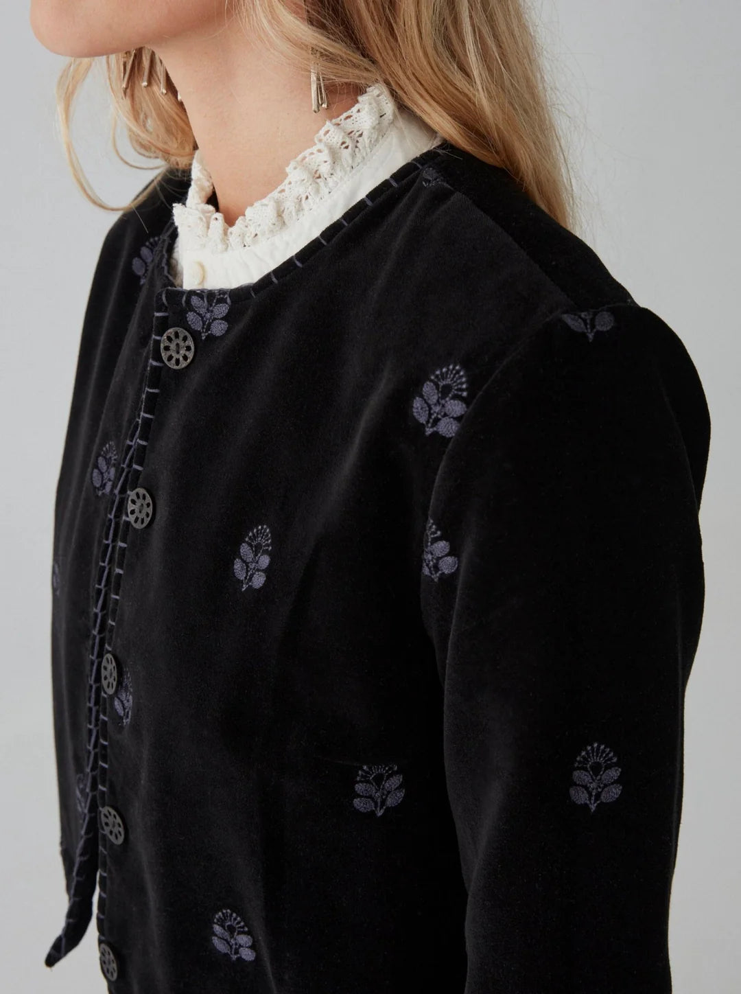 A person's upper body, featuring a black embroidered jacket with floral patterns and a white lace collar underneath.