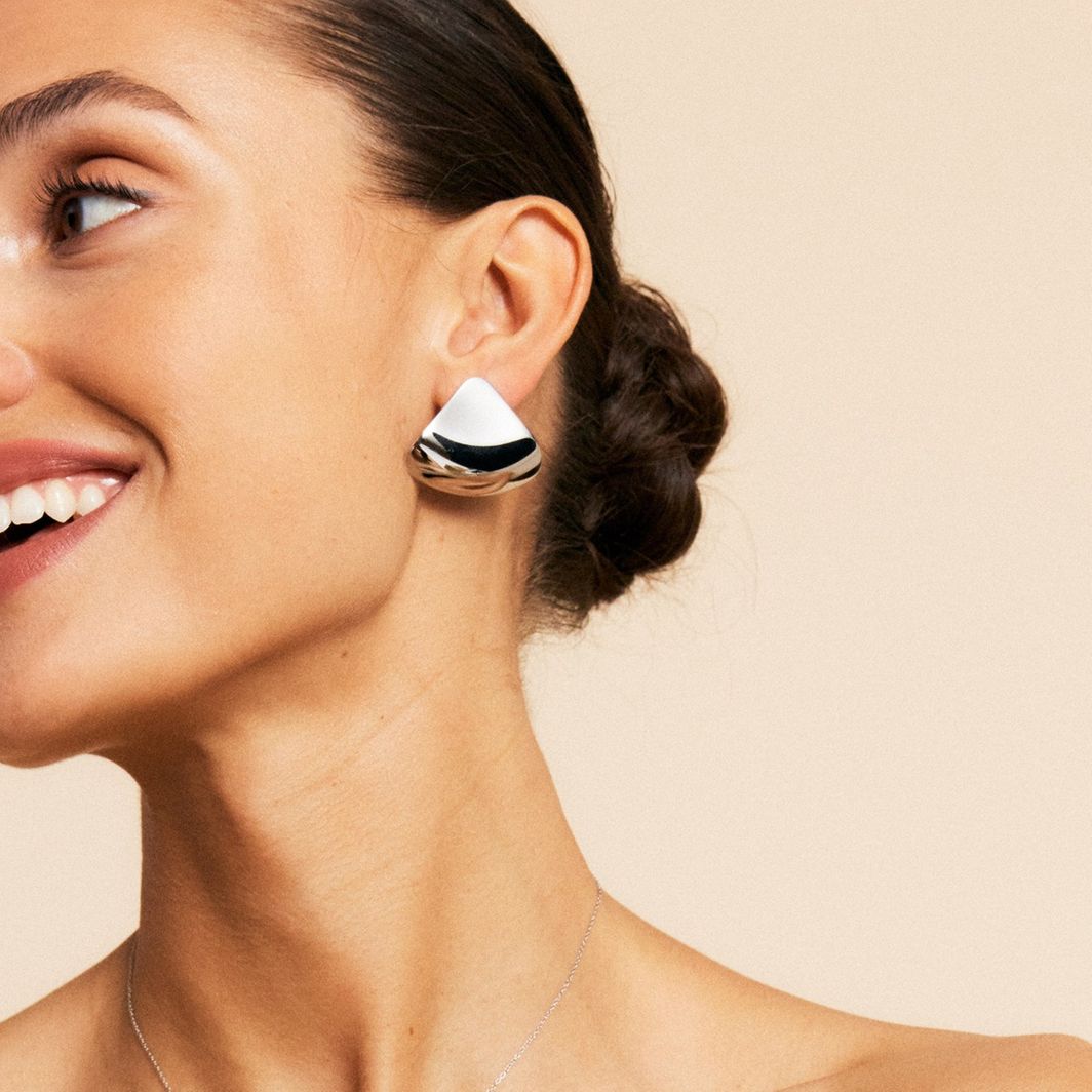 A person with swept-back hair and a smile wears a large geometric earring. The background is a neutral beige.