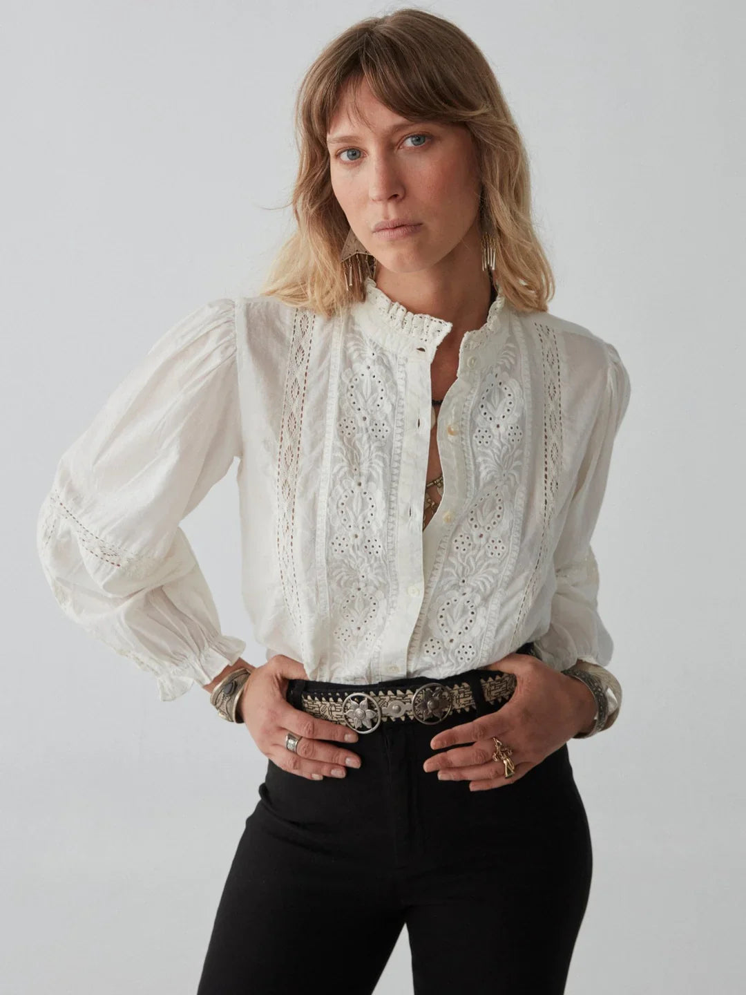 A person wearing a white embroidered blouse and black pants poses against a plain background. They have shoulder-length hair and wear bracelets and rings.