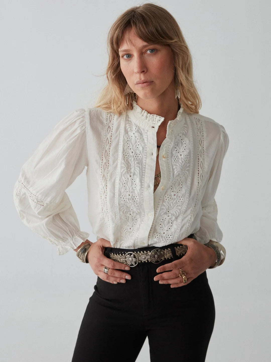 A person with medium-length hair wears a white, long-sleeve blouse with lace details and black high-waisted pants. They have multiple bracelets and rings and stand against a plain background.