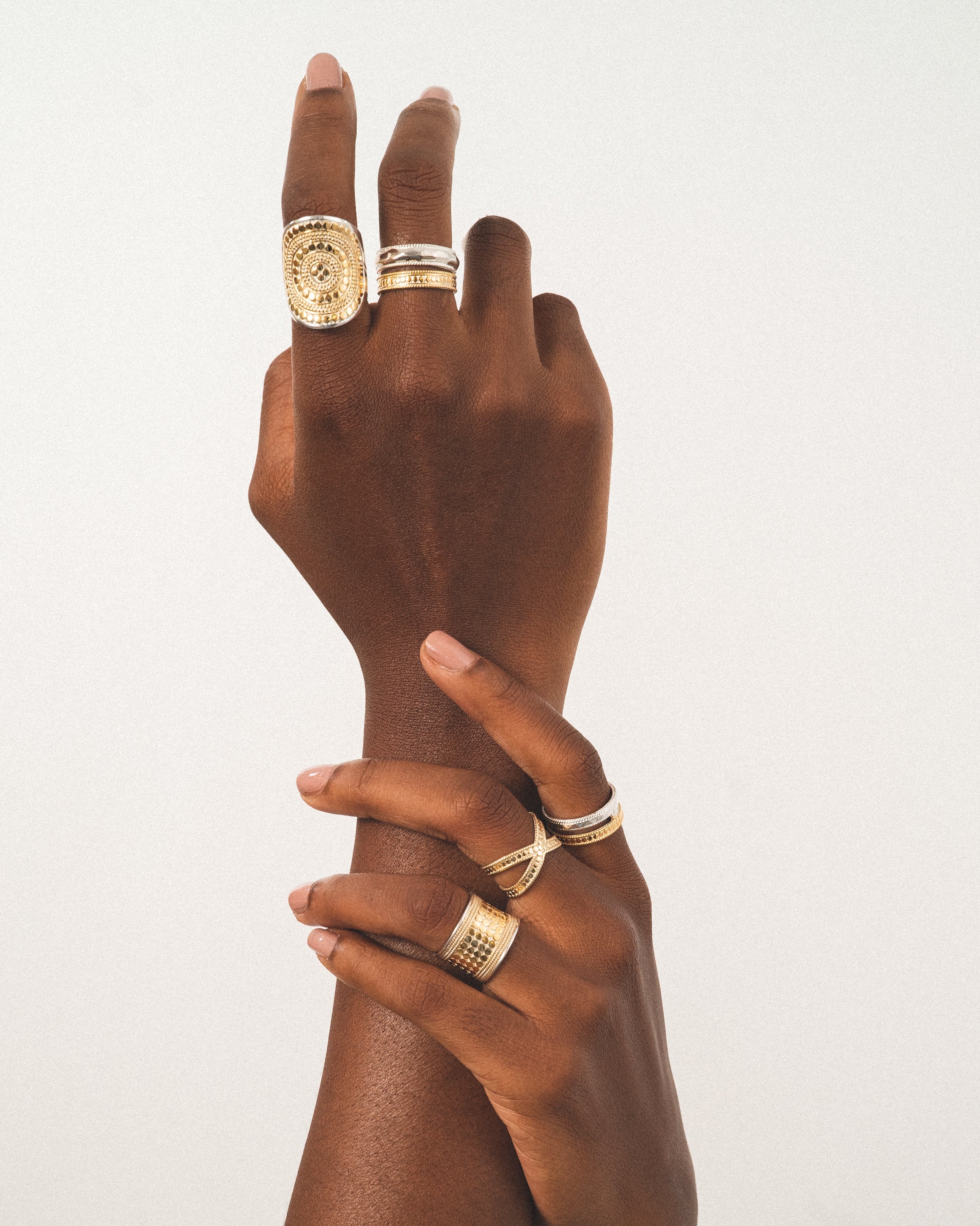 Two hands adorned with various rings in gold and silver are positioned against a neutral background.