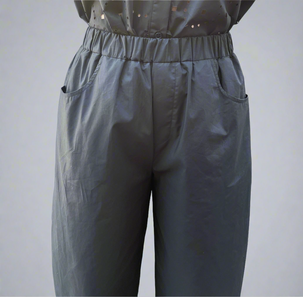 Someone is wearing MeiMeij's Barrel Leg Cotton Trousers in black, featuring a loose fit, elastic waist, and side pockets. The silky-soft look complements the simple backdrop of a white wall with a blue stripe.