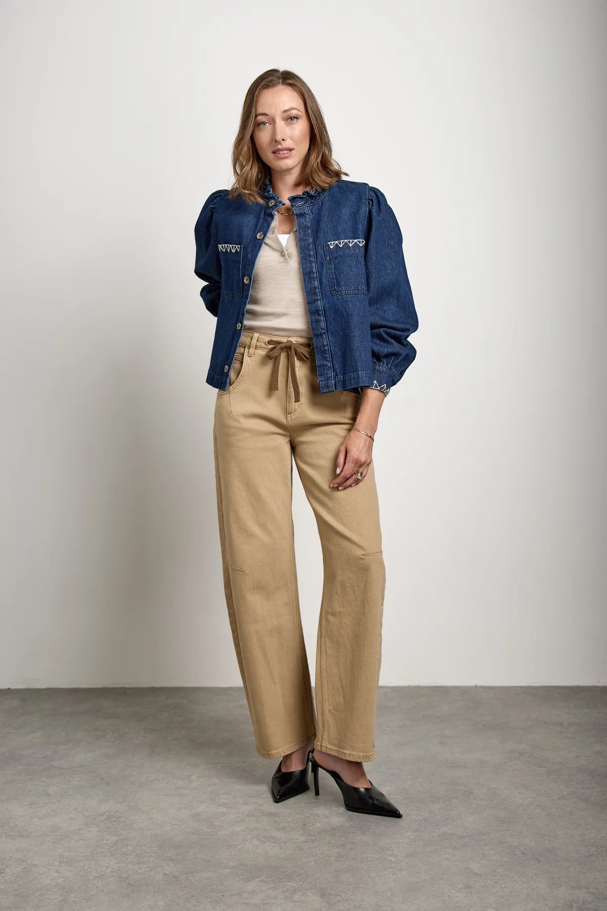 A woman stands indoors against a plain background, wearing the Seventy & Mochi Ziggy Jacket in Americana—a blue relaxed fit denim jacket—styled with a light beige top, tan high-waisted organic cotton trousers with a tie belt, and black heeled shoes.