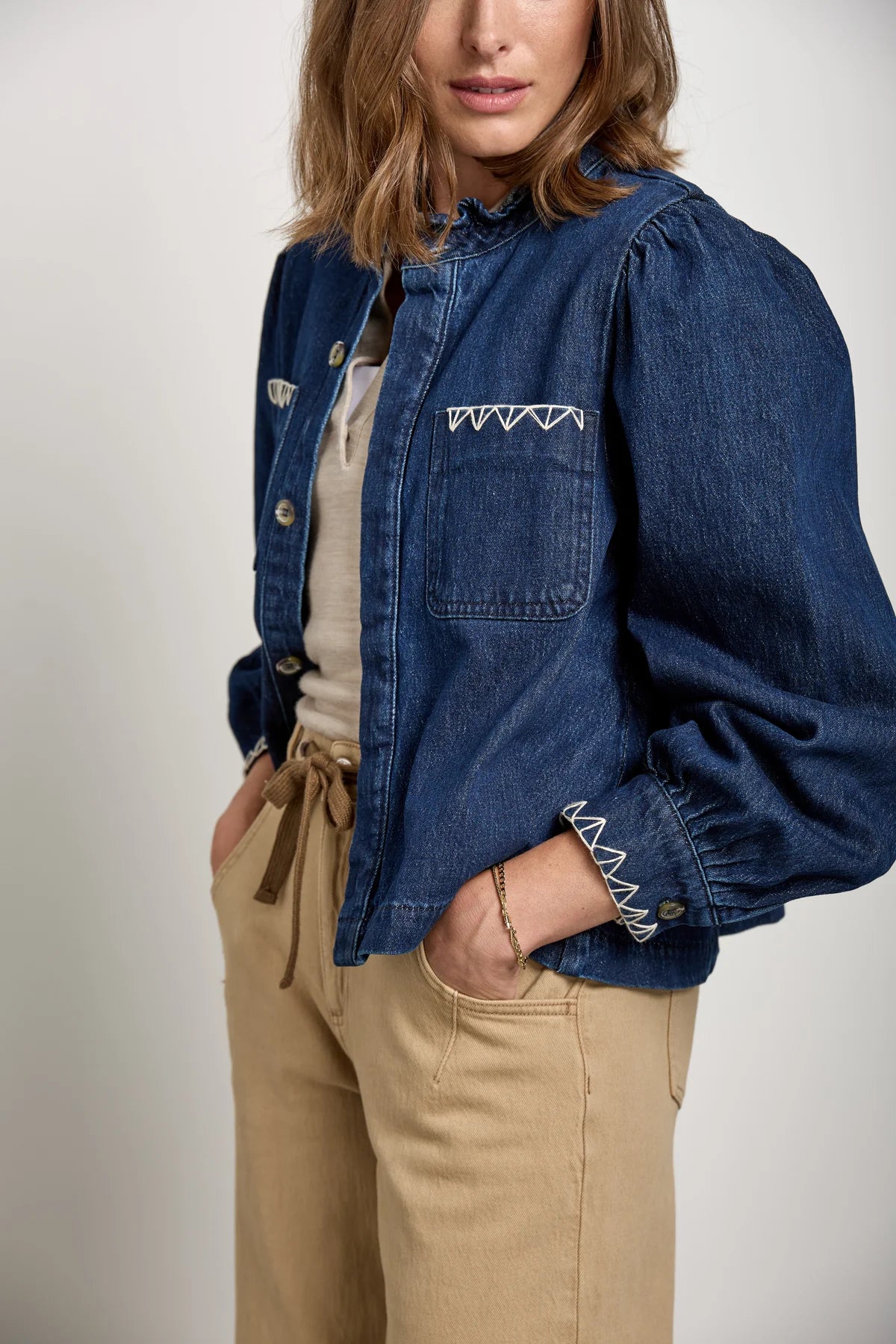 A woman stands with her hands in her pockets against a plain background, wearing the Seventy & Mochi Ziggy Jacket - Americana, a relaxed fit blue denim jacket with white stitching, paired with beige pants.