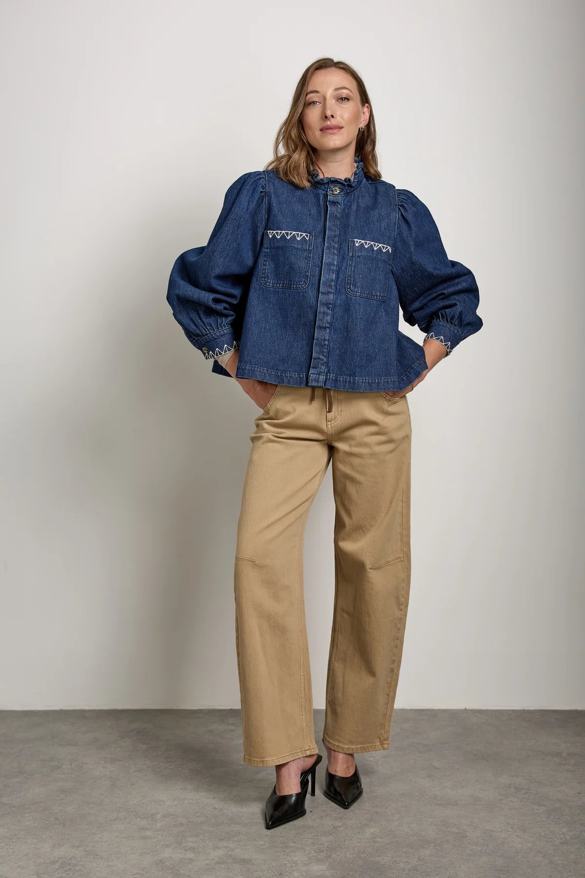 A woman stands against a plain wall wearing the Seventy & Mochi Ziggy Jacket in Americana, styled with beige wide-leg pants and black heeled shoes, her hands in her pockets.
