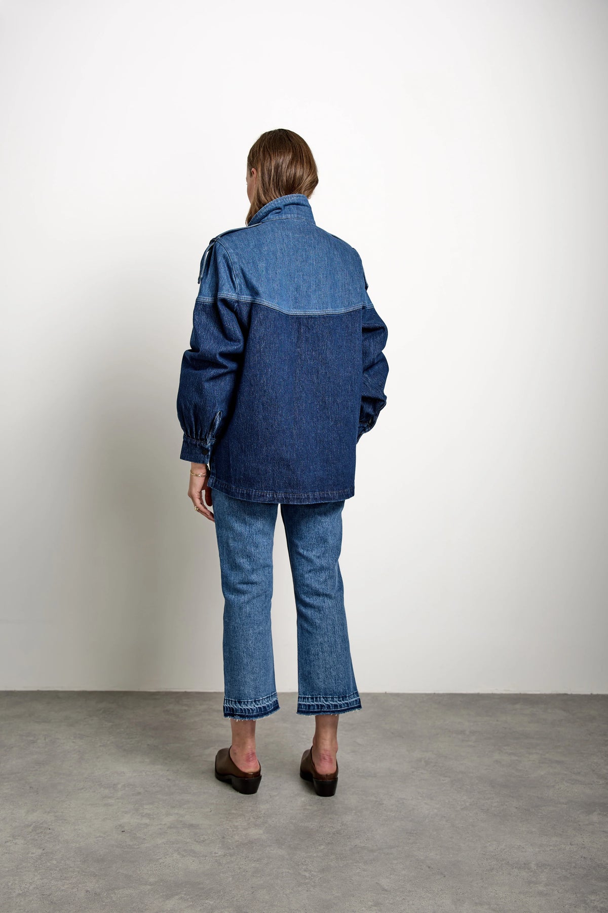 A person stands indoors facing away, wearing the Seventy & Mochi Bowie Jacket - Americana paired with blue jeans and brown shoes, short hair visible. The background is plain white with a gray floor.