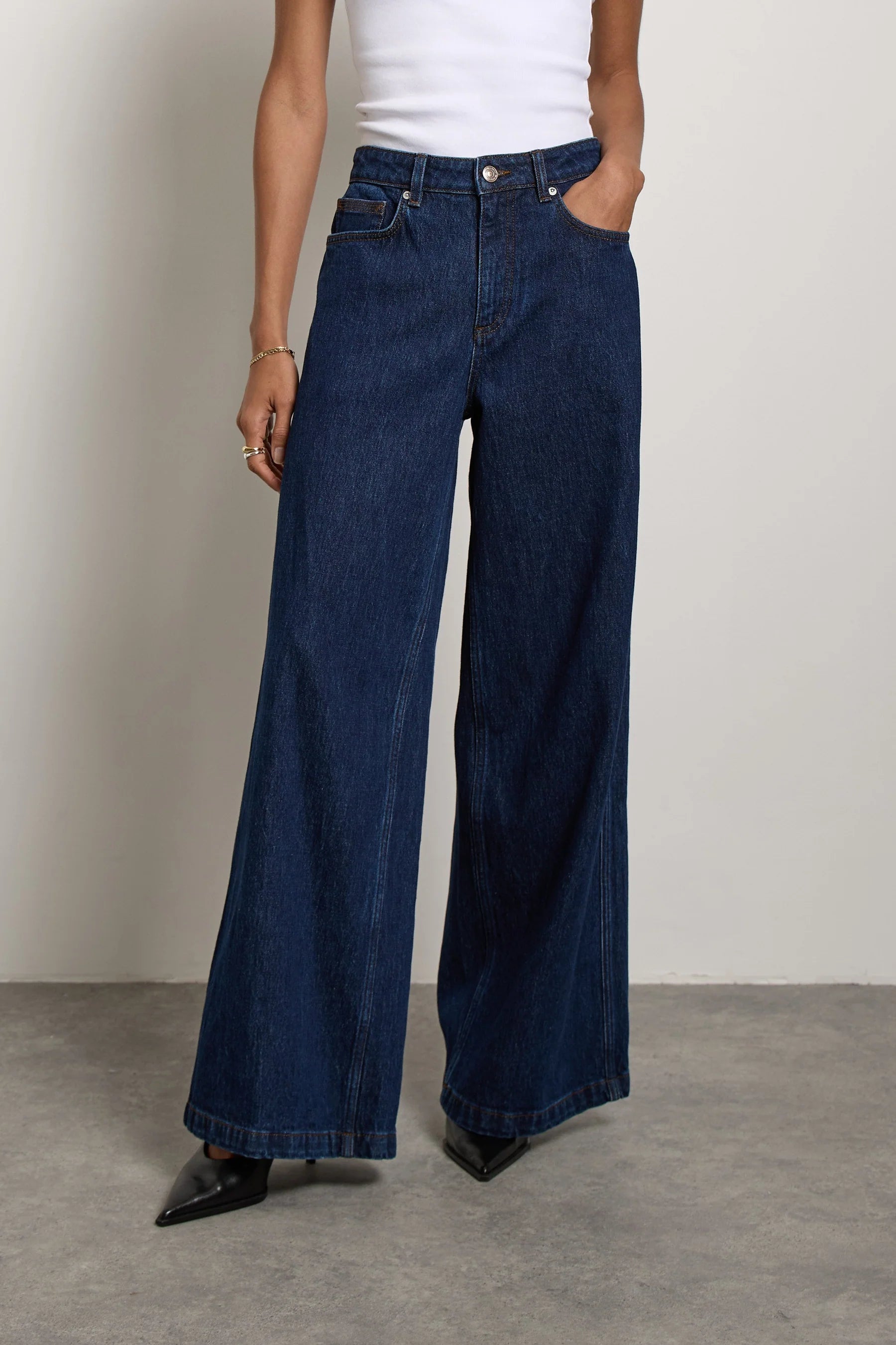 A person wearing a white sleeveless top and Seventy & Mochi’s Bay jean in Americana—super high rise, extra-wide leg dark blue rigid denim—with one hand in a pocket and black pointed shoes visible.