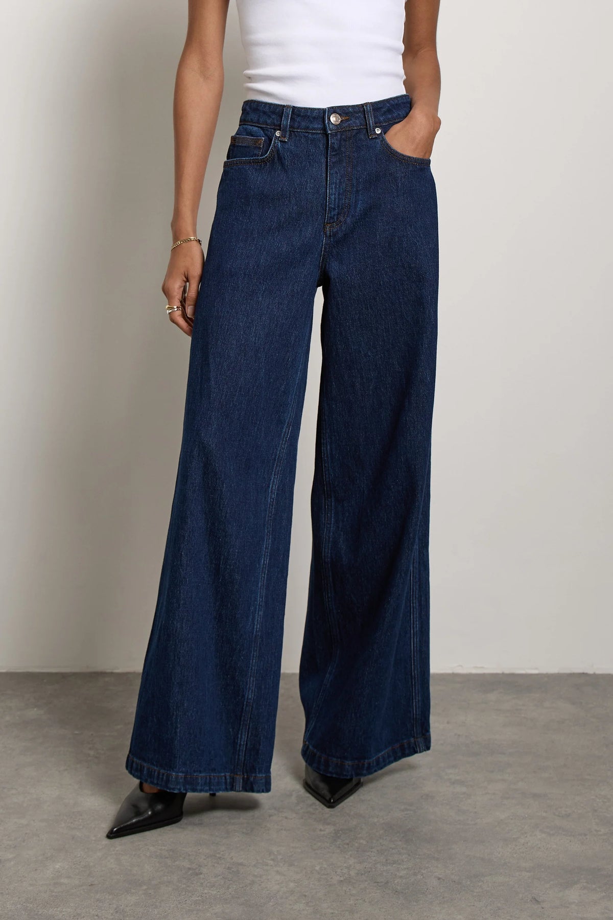 A person wearing a white sleeveless top and Seventy & Mochi’s Bay jean in Americana—super high rise, extra-wide leg dark blue rigid denim—with one hand in a pocket and black pointed shoes visible.