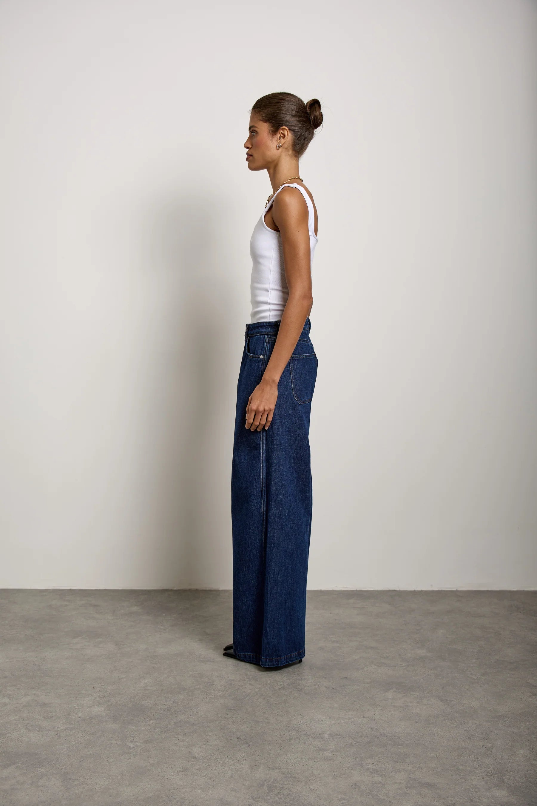 A woman in profile wears a white tank top and the Bay jean in Americana by Seventy & Mochi, featuring a super high rise and extra-wide leg, standing against a plain off-white wall on a gray floor.