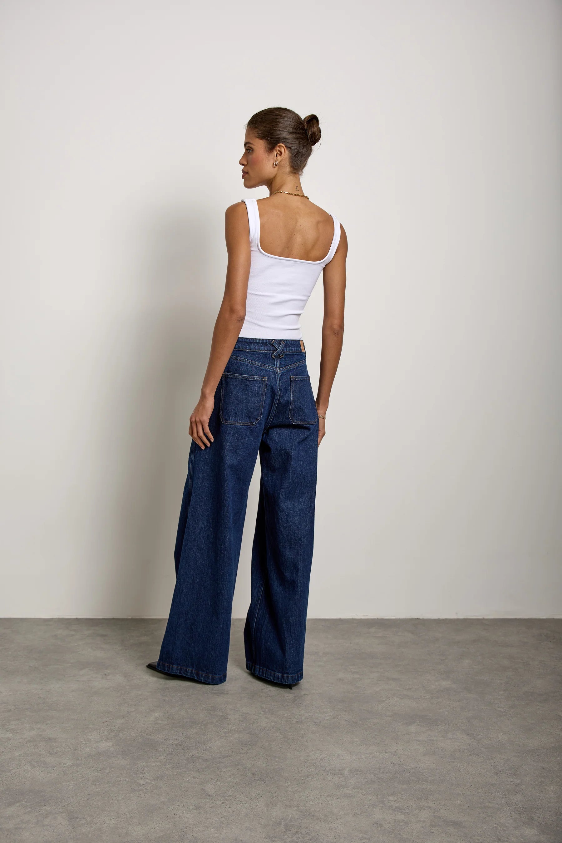 A woman with her hair tied back in a bun stands on a gray floor facing away from the camera, wearing Seventy & Mochi’s Bay jean in Americana—extra-wide leg, rigid denim jeans—paired with a white sleeveless top. The backdrop is a plain off-white wall.