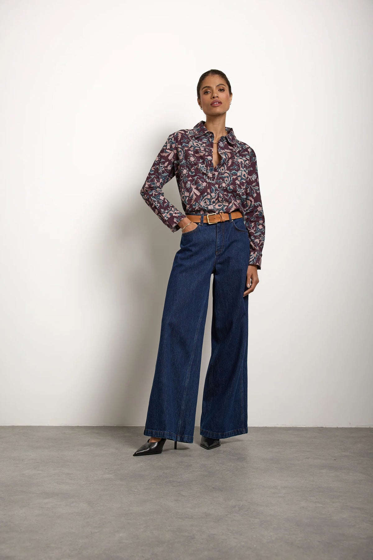 A person stands against a white wall wearing a patterned long-sleeve shirt, the Bay jean in Americana by Seventy & Mochi—a pair of super high-rise, rigid denim extra-wide leg jeans—styled with a brown belt and black pointed-toe shoes.