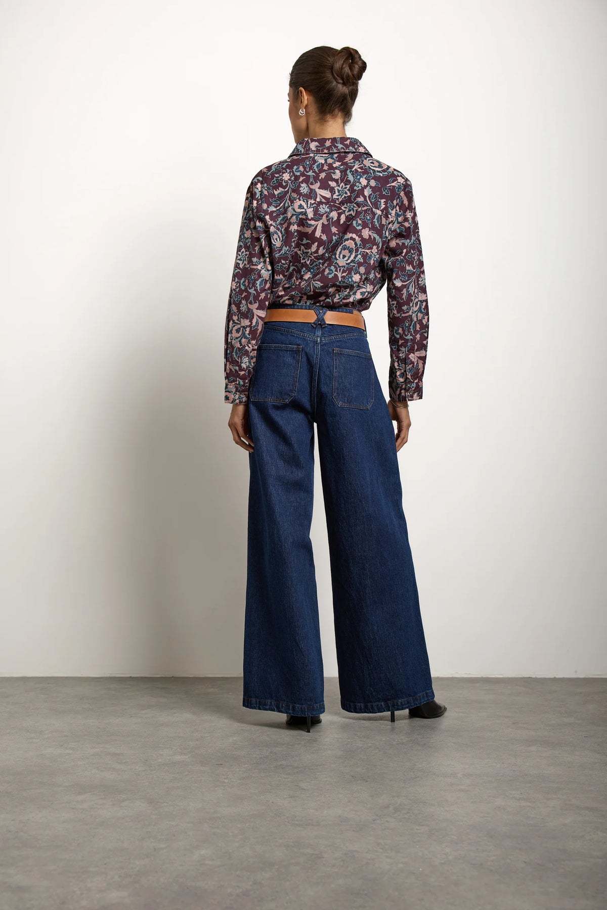 A person standing indoors with their back to the camera, wearing a patterned long-sleeve shirt, black heeled shoes, hair styled in a bun, and the Bay jean in Americana by Seventy & Mochi—super high rise extra-wide leg jeans in rigid denim.