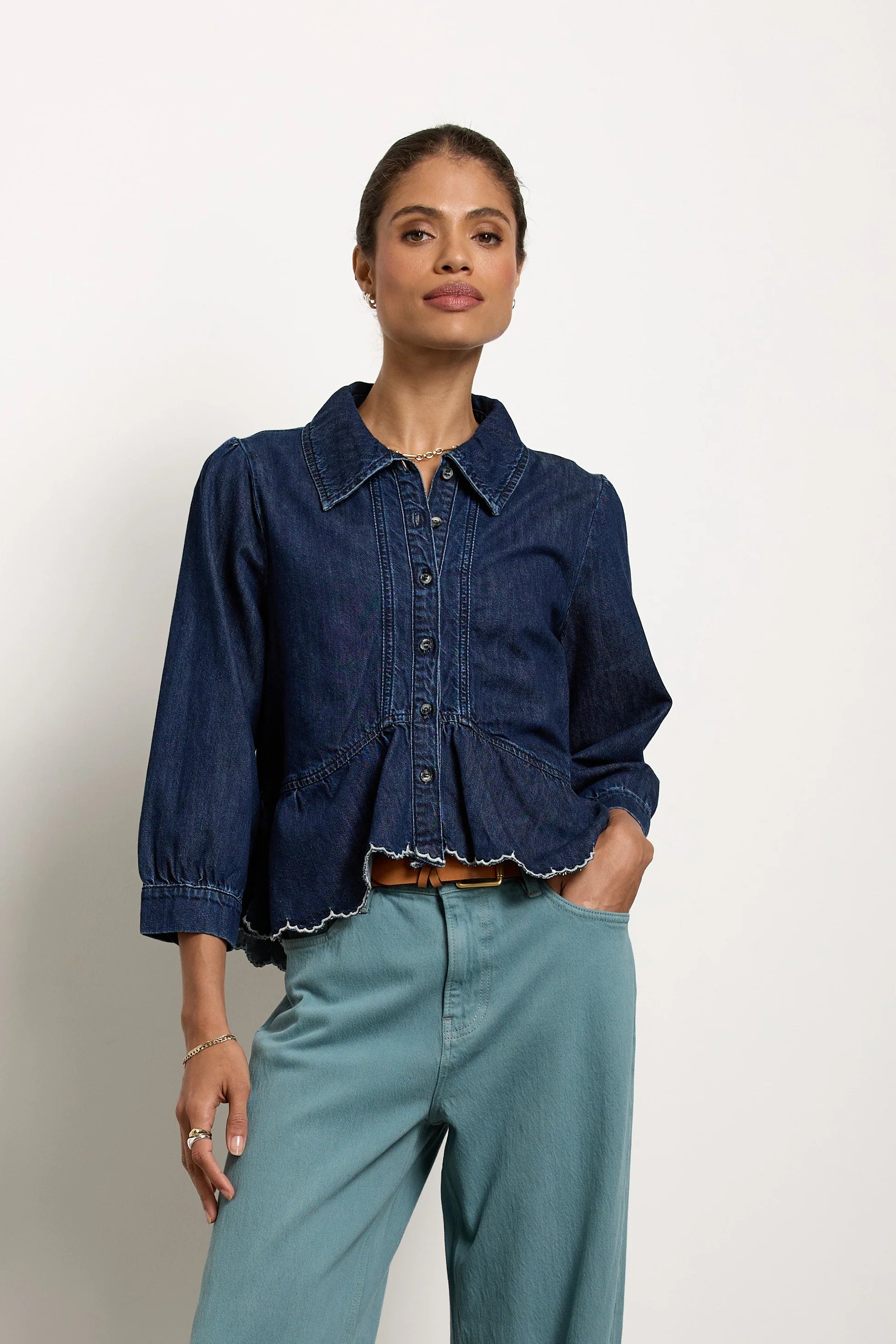 A model stands against a white wall wearing a dark blue denim shirt tucked into light blue wide-leg trousers, paired with a brown belt and dark brown shoes.