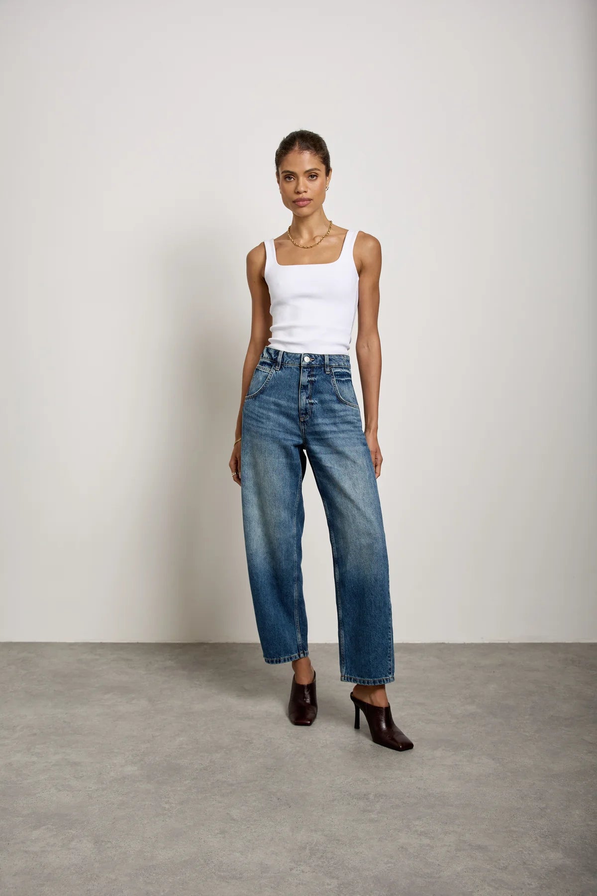 A woman stands against a plain wall wearing a white tank top, Seventy & Mochi’s Bo Barrel Leg Jeans in Phoenix crafted from rigid denim, brown heeled boots, and a necklace.