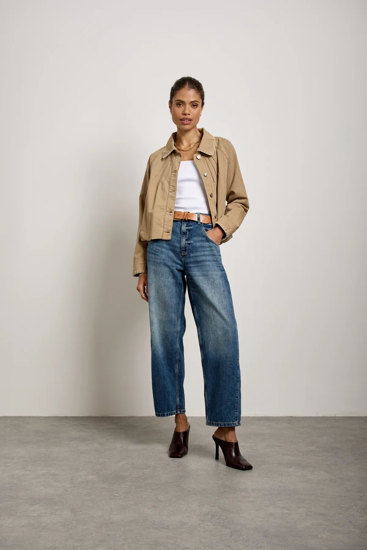 A woman stands against a plain wall wearing a beige jacket, white crop top, and Seventy & Mochi’s Bo Barrel Leg Jeans in Phoenix—a pair of blue super high-rise jeans crafted from rigid denim—paired with brown heeled boots.