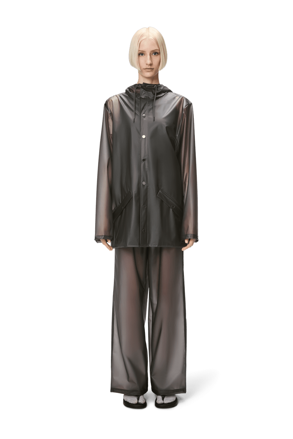 A person is elegantly dressed in Rains' lightweight, transparent gray Jacket W3 and matching pants against a plain background, blending unisex style and practicality for any weather.