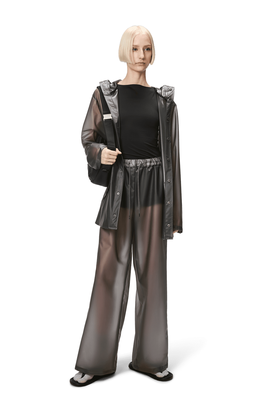 A person stands in a transparent Rains Jacket W3 and matching pants over a black outfit, with a neutral expression. The unisex ensemble appears lightweight, offering both style and functionality.