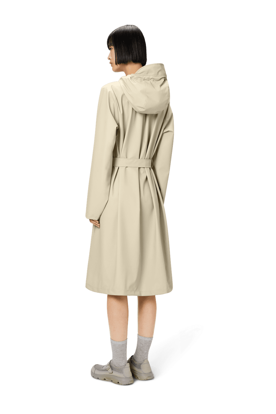 A person with short hair stands facing away, wearing Rains' A-Line Longer W Jacket in beige, made from waterproof PU fabric and featuring a hood and belt. They pair it with gray socks and matching beige shoes to create an elegant silhouette.