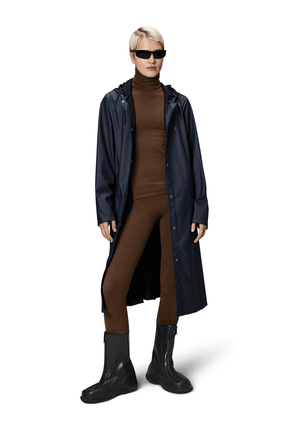 A person stands wearing sunglasses, a brown turtleneck, matching pants, black boots, and the A-Line Longer W Jacket by Rains, a long dark raincoat made from PU fabric.