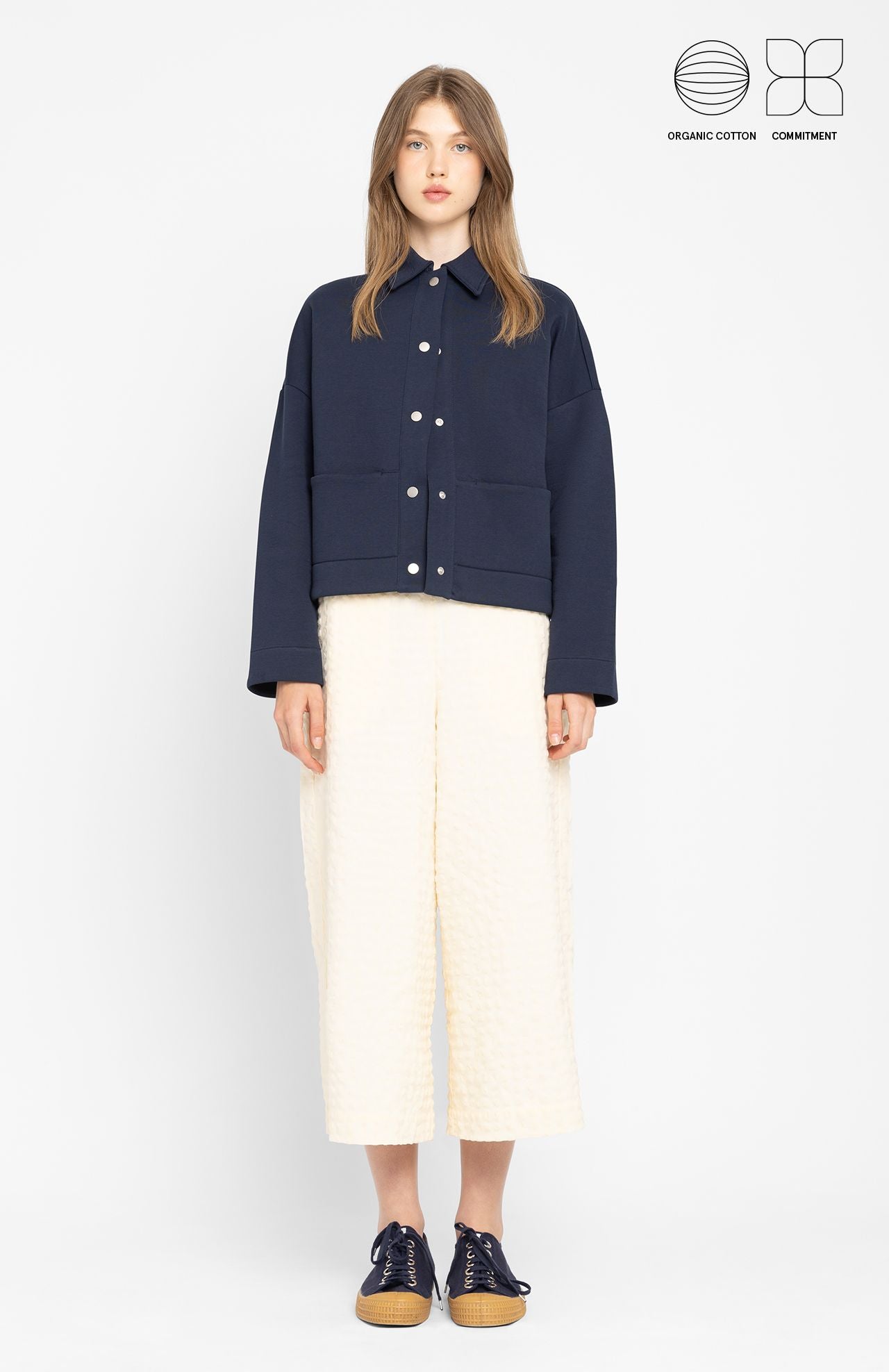 A person in a Loreak Mendian Ixiar Jacket - Navy, paired with cream textured pants, stands against a white background. Icons highlight the spongy fabric, emphasis on organic cotton, and commitment.