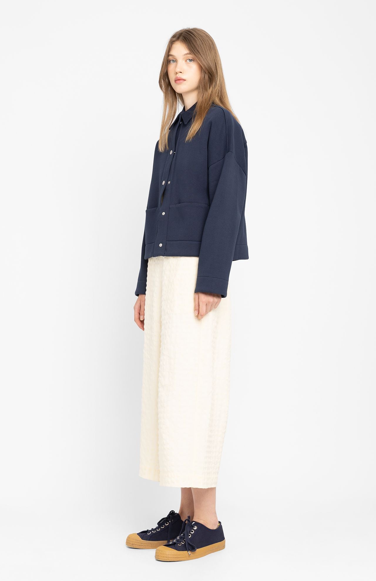 A person stands against a plain white background, wearing a Loreak Mendian Ixiar Jacket - Navy, paired with a white skirt and navy sneakers with tan soles.