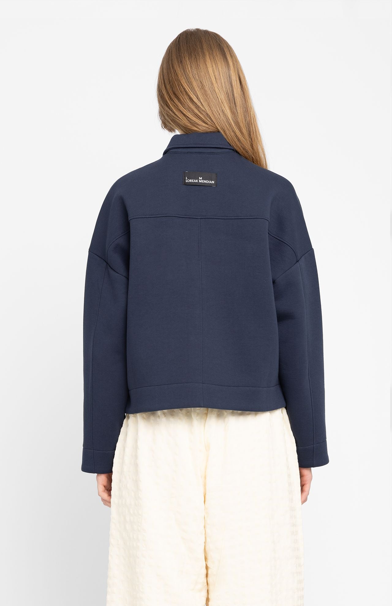 A person with long hair, wearing a Loreak Mendian Ixiar Jacket - Navy over a white skirt, stands facing away from the camera. This spongy fabric jacket is a versatile staple that effortlessly elevates their style.