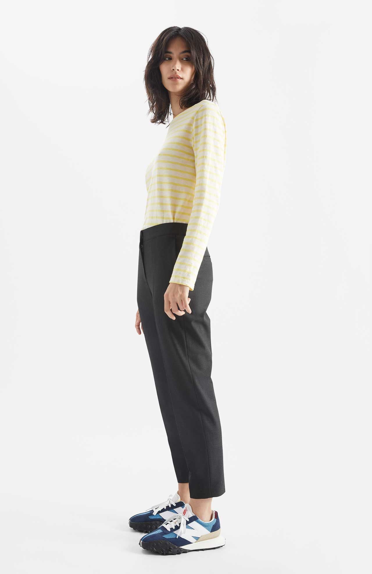 Person wears Alaiak trousers in a dark grey shade. The pants are worn with a yellow and white striped long sleeve top. Side view.