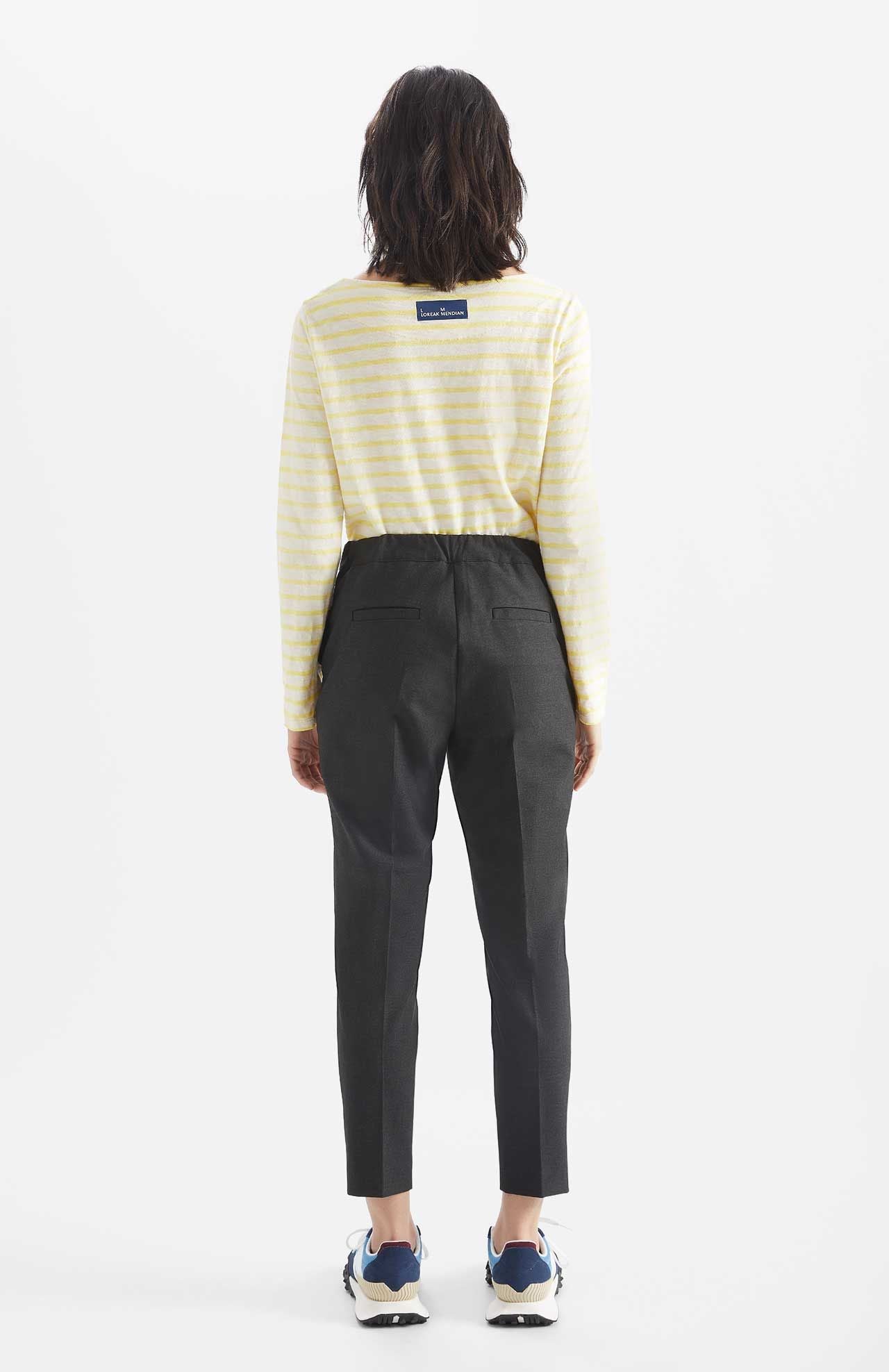 Person wears Alaiak trousers in a dark grey shade. The pants are worn with a yellow and white striped long sleeve top. Back view.