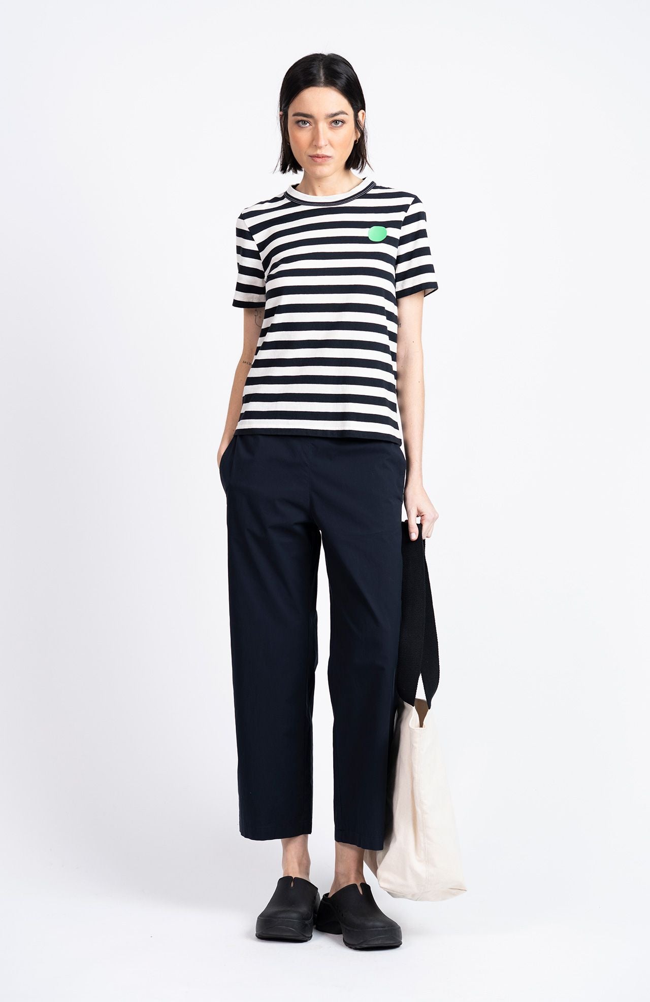 A woman stands facing forward wearing a black and white striped t-shirt, Loreak Mendian Confya Cropped Trousers in navy, black shoes, and holding a cream tote bag with a black handle.