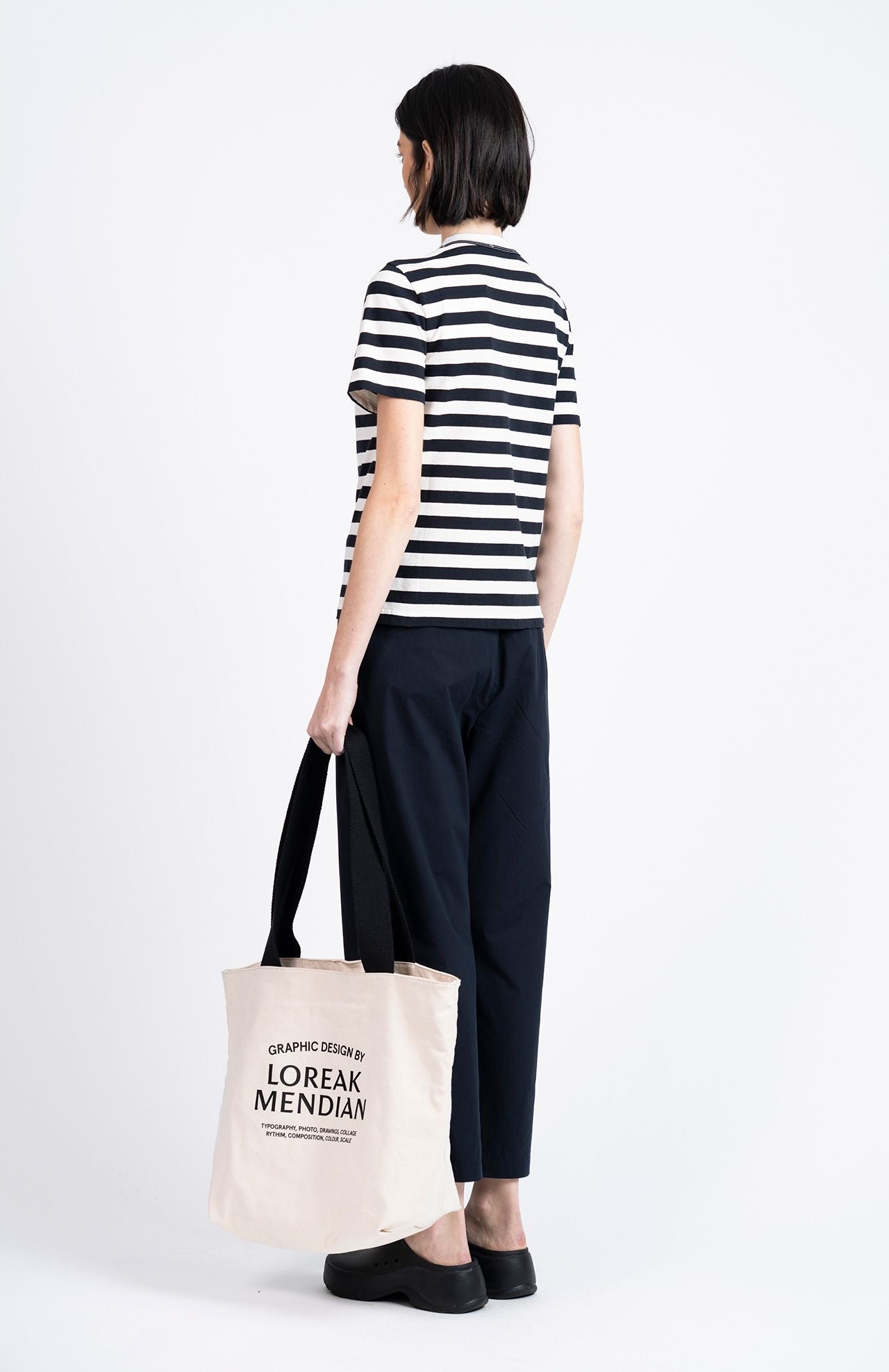 A person stands facing away against a white background, wearing a black and white striped t-shirt, Loreak Mendian Confya Cropped Trousers in navy, black shoes, and holding a white text-printed tote bag.
