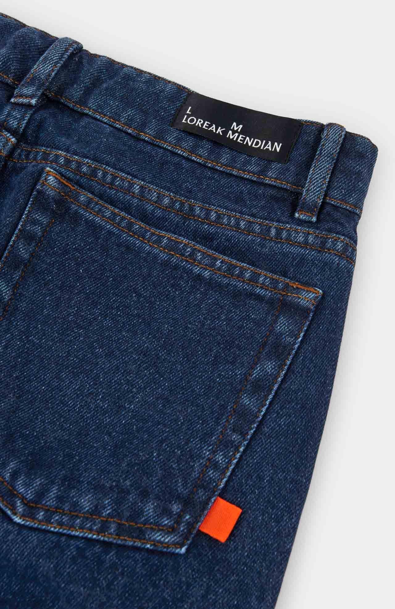 Close-up of the back pocket on Navasota Denim Trousers - Indigo by Loreak Mendian, featuring a black brand label and a small orange fabric tag.