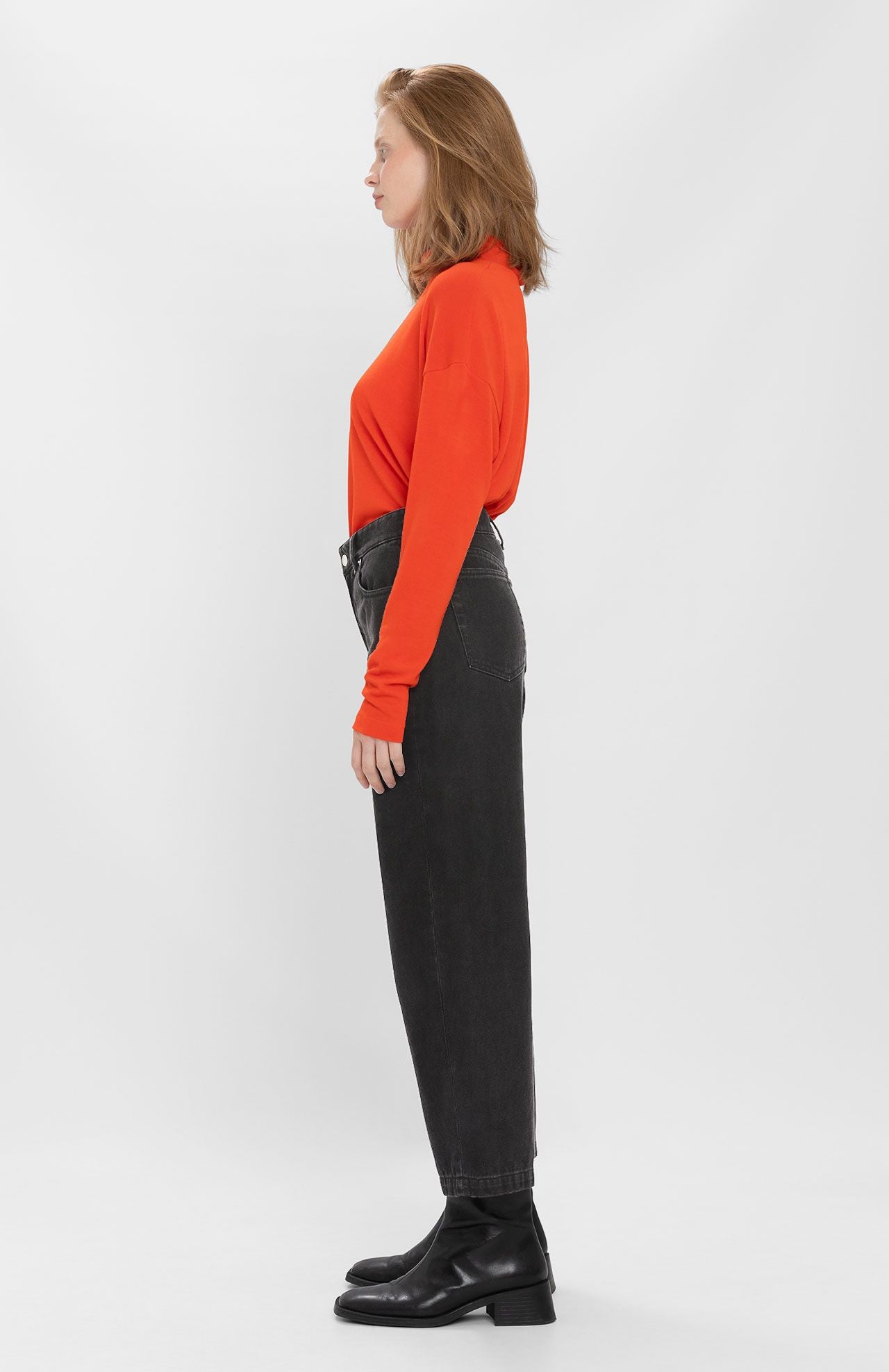 A person in a side profile poses against a white background, wearing a long-sleeve red top, high-waisted Navasota Denim Trousers - Black from Loreak Mendian made from organic cotton, and black boots.