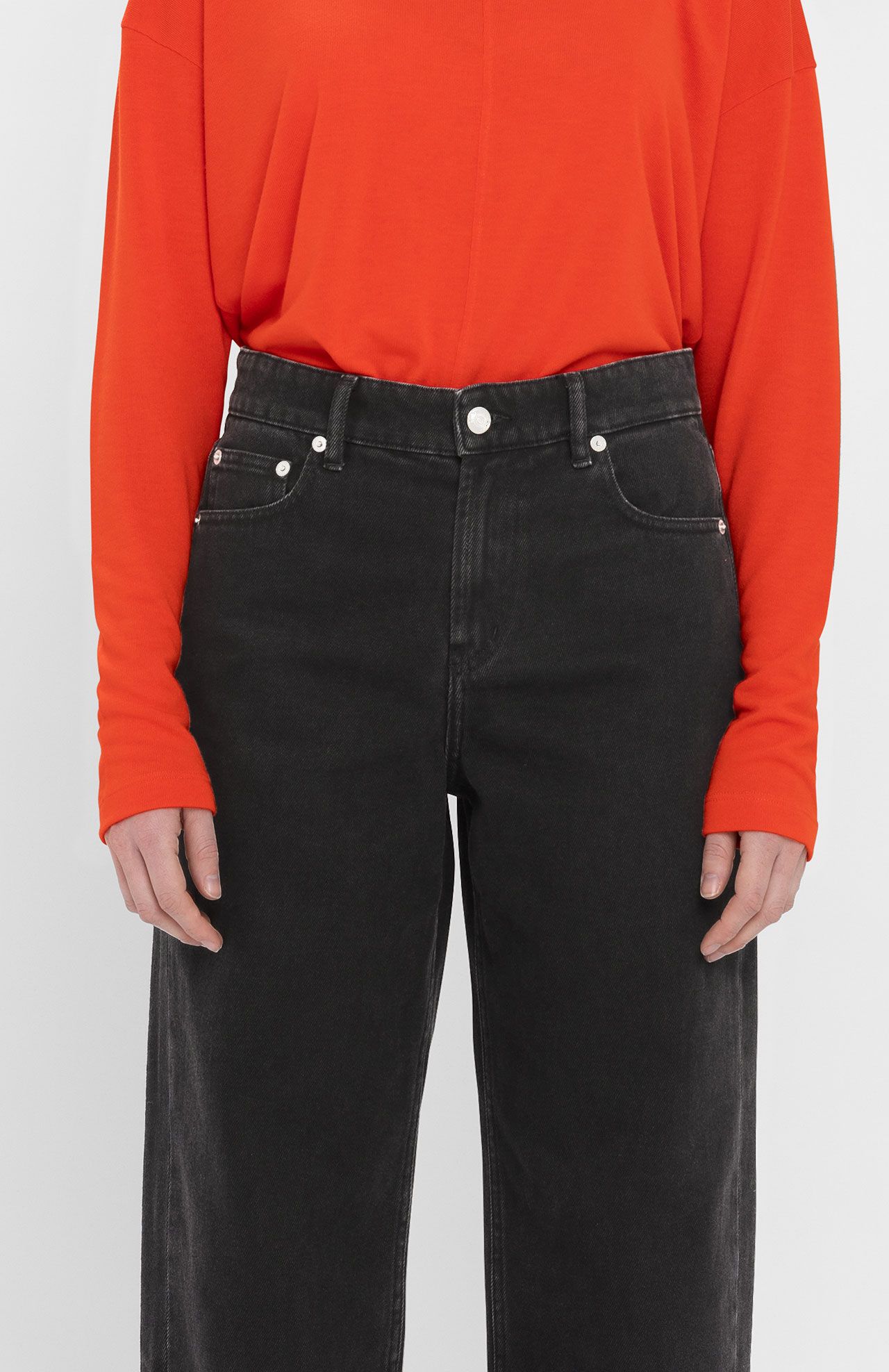 A person wearing a bright orange long-sleeve shirt and high-waisted Navasota Denim Trousers - Black from Loreak Mendian, made from organic cotton, stands against a plain background.