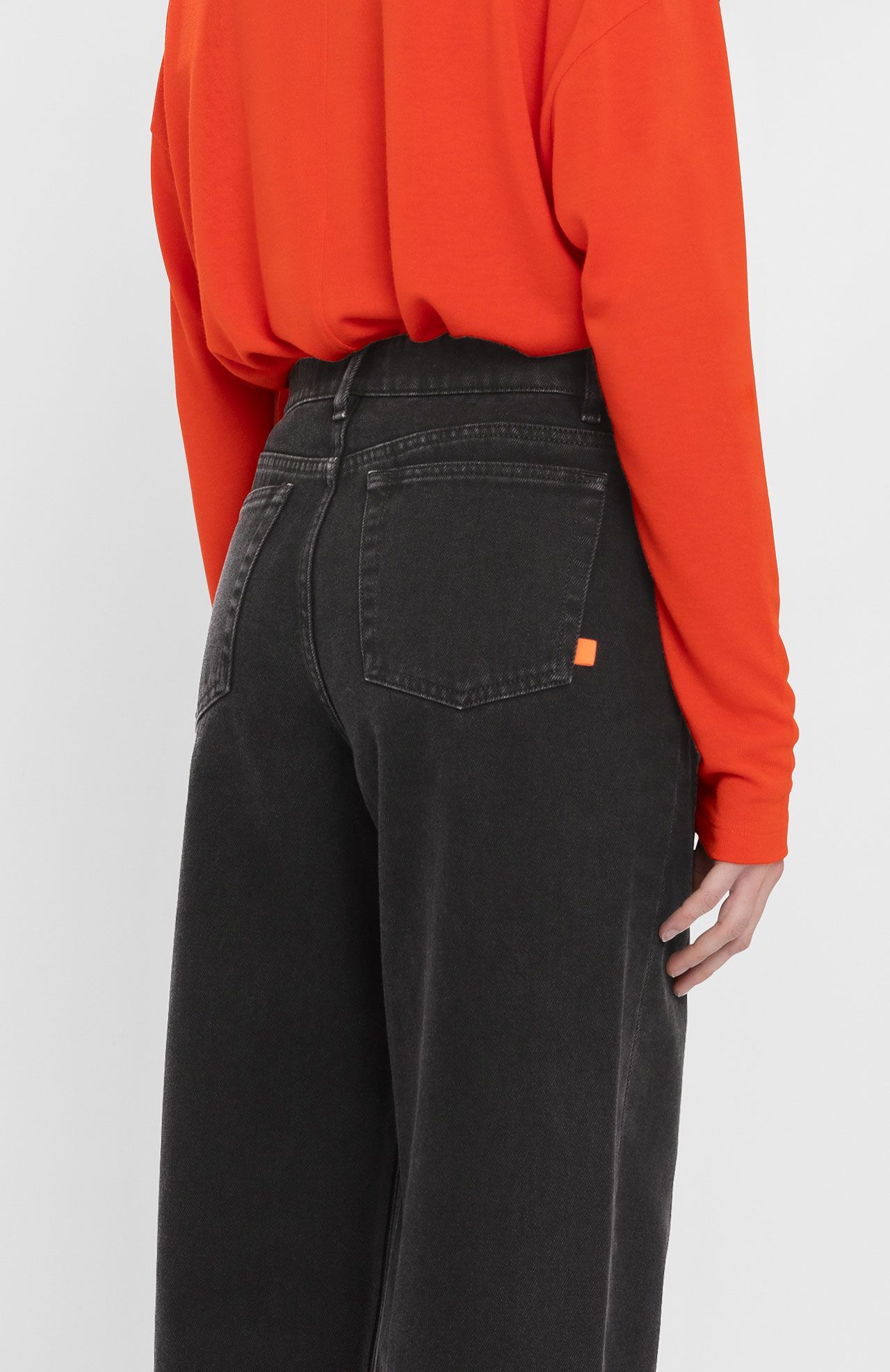 A person wearing a loose-fitting, bright orange top and Loreak Mendian's high-waisted black Navasota Denim Trousers made from organic cotton is seen from the back. Their right arm is slightly bent, and a small orange tag is visible on the jeans pocket.